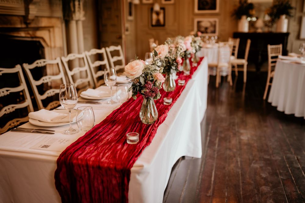 Elegant wedding table setting with white linens, red velvet runner and floral centerpieces in an ornate dining room.