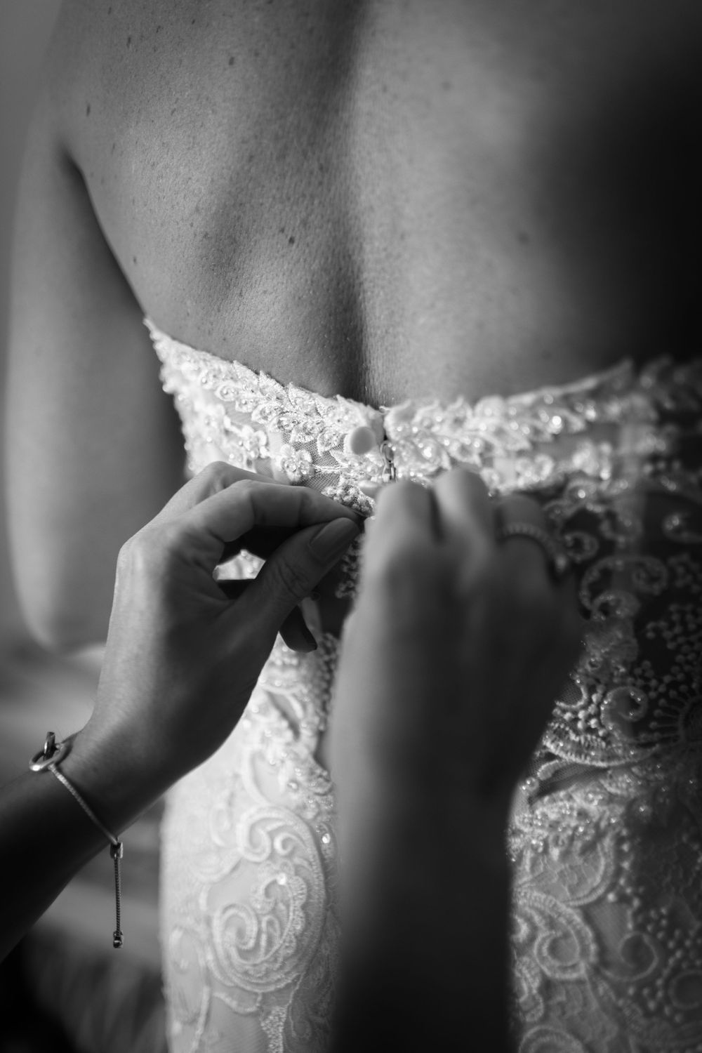 Close-up black and white detail of hands fastening delicate lace on a wedding dress bodice.