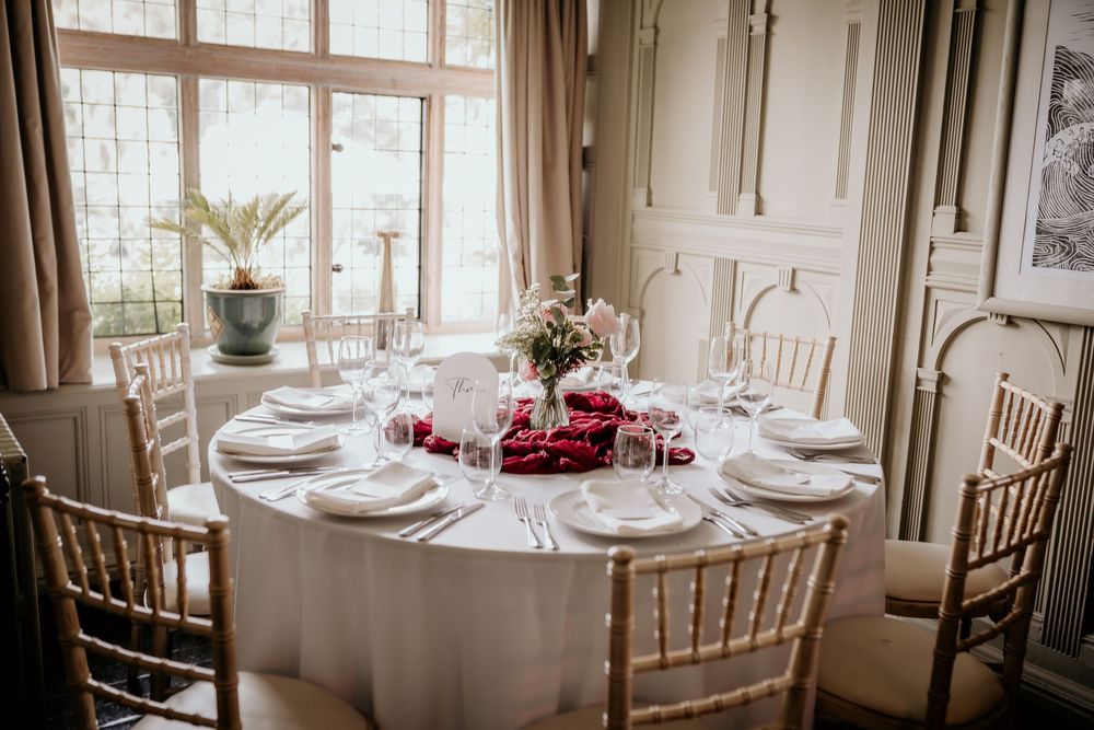 Elegant round dining table with white linens and chiavari chairs set for a formal event near bright windows.