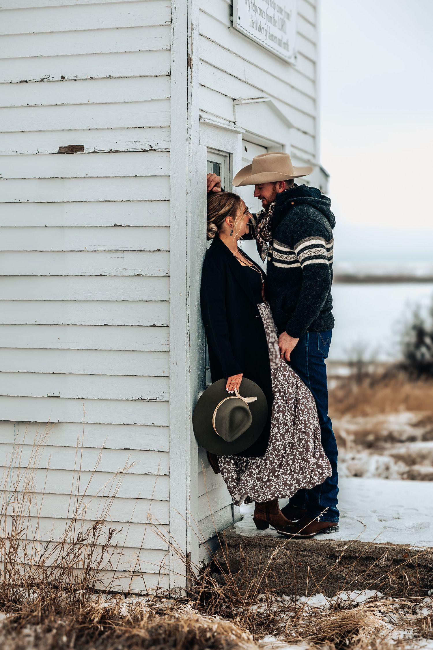 A romantic western couple embraces beside a white wooden building during winter, in Drumheller, Alberta.