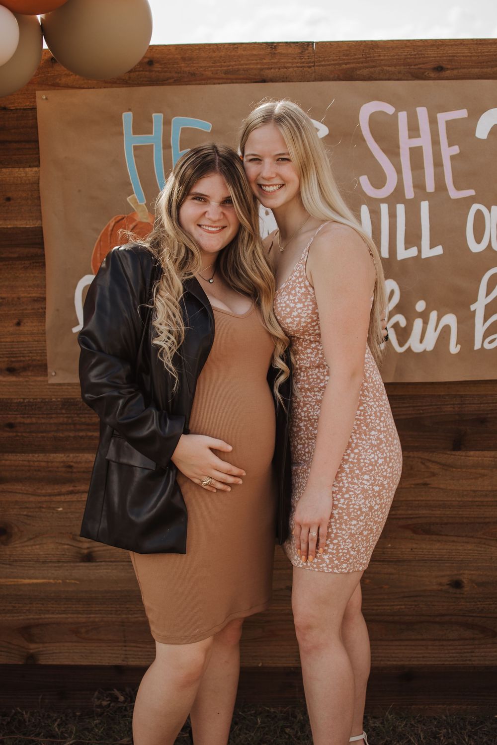 Two people posing together against a wooden wall with text, one wearing a black jacket and tan dress, another in pink sequins.