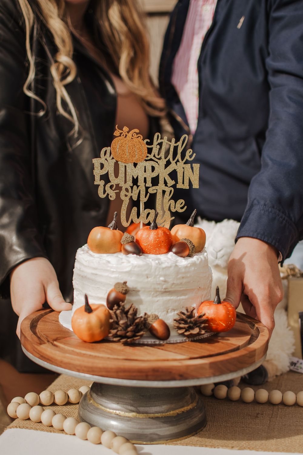 A white frosted cake decorated with small orange pumpkins and pinecones on a wooden cake stand with a gold topper.