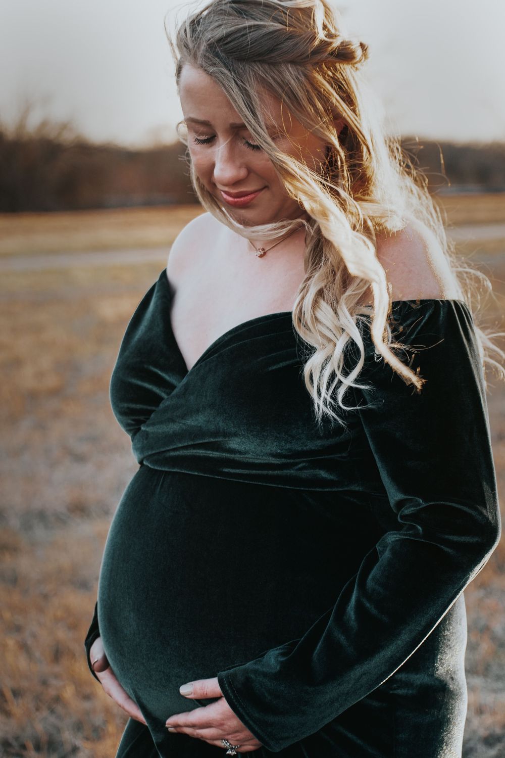 Expectant mother in dark green velvet dress touches her baby bump while smiling during outdoor golden hour portrait session.