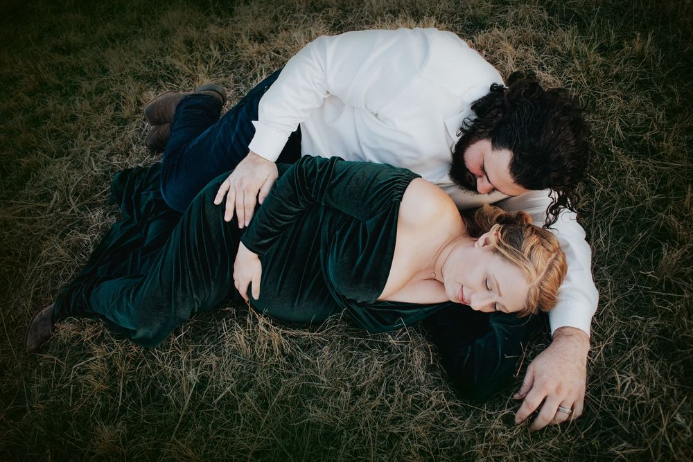 A romantic couple lying together on grass wearing formal attire in dark green and white during a photoshoot.