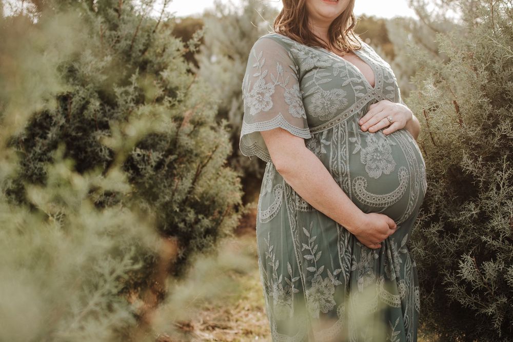 Expecting mother in sage green lace maternity dress poses outdoors among foliage during golden hour photoshoot.