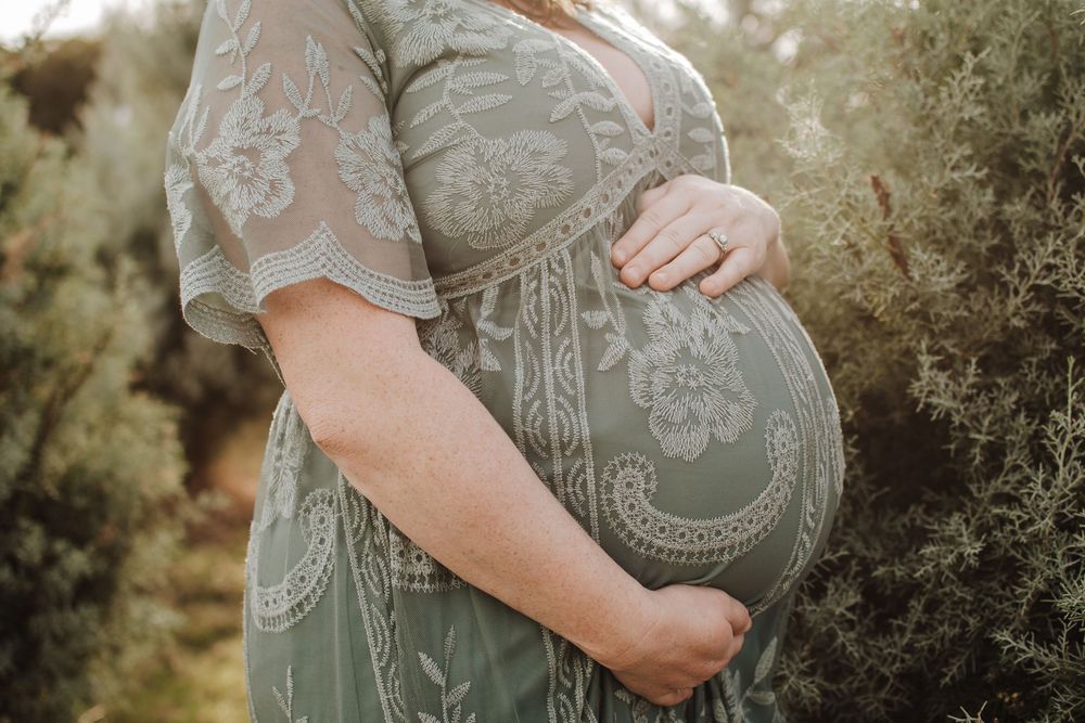 A pregnant person in a sage green lace dress cradles their belly during a golden hour maternity photoshoot outdoors.