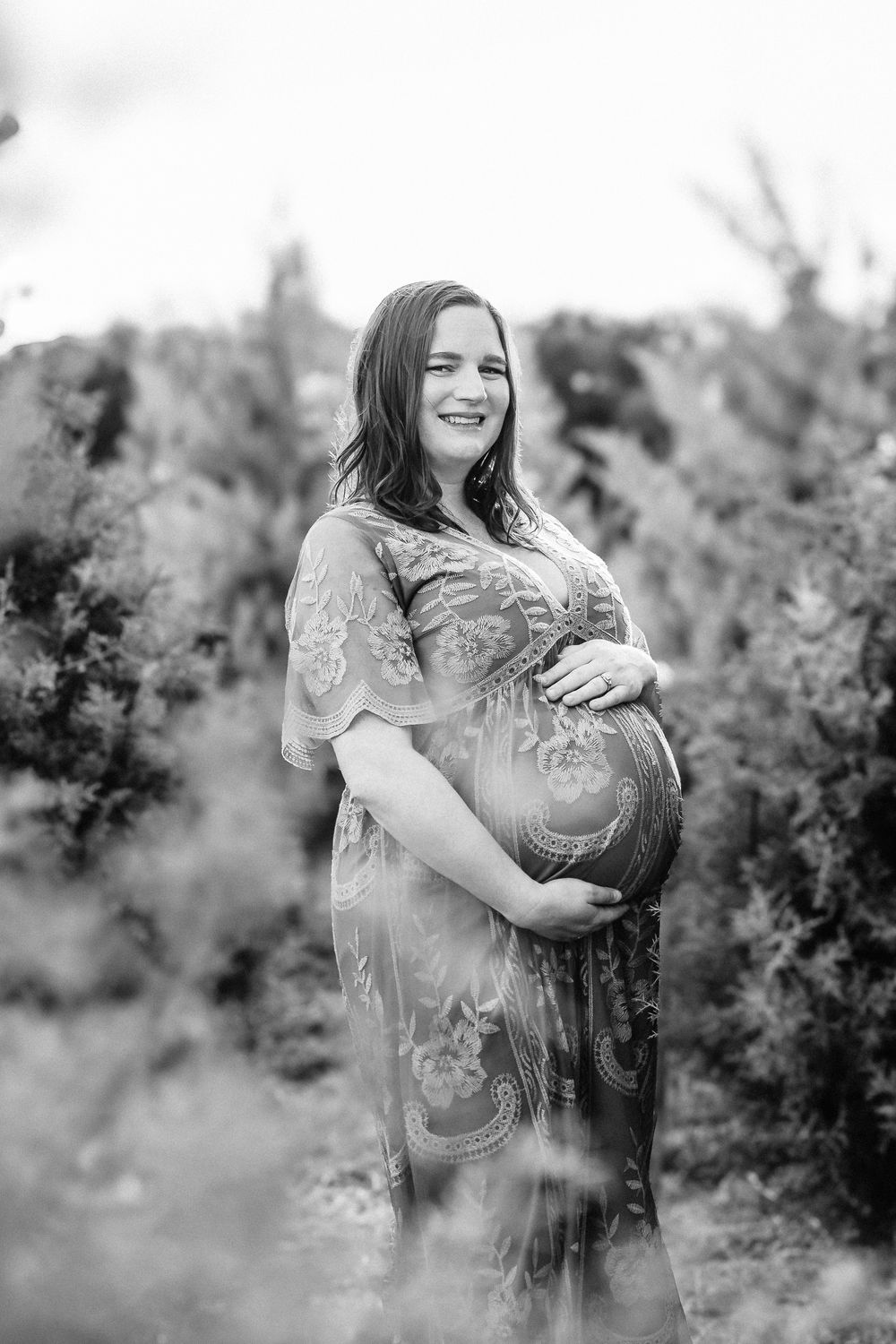 A pregnant woman in a floral dress poses outdoors during a black and white maternity photo shoot among greenery.
