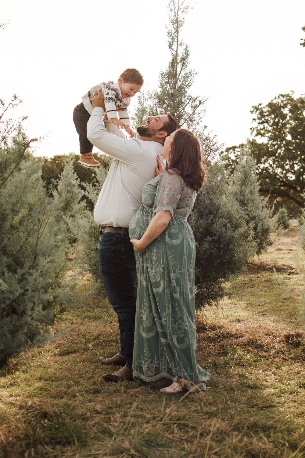 A couple shares a tender moment outdoors lifting their small child against an evergreen backdrop at sunset.