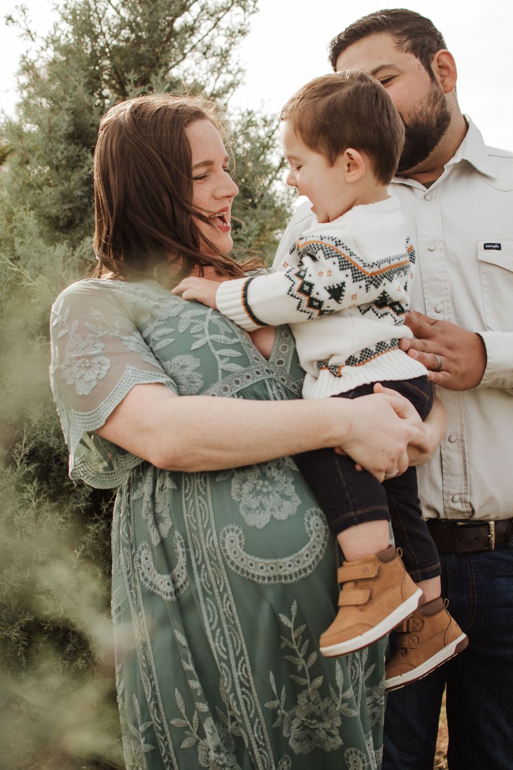 Family shares a tender moment outdoors with vegetation in background as they embrace and smile together.