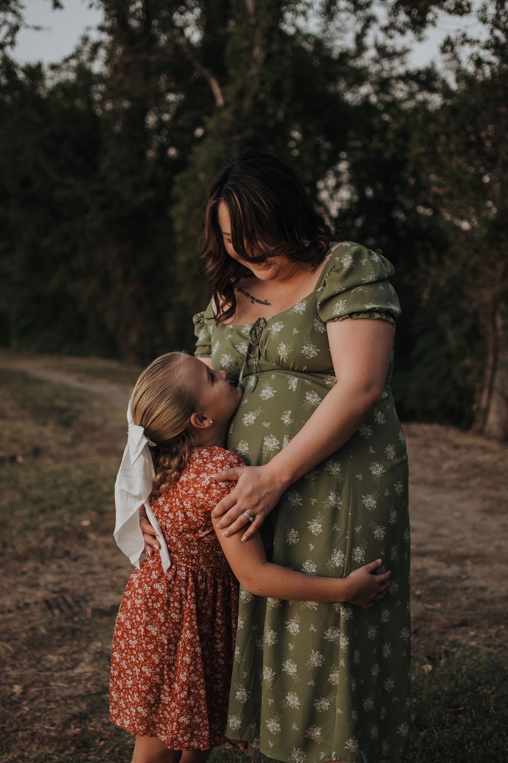 A tender outdoor moment between parent and child wearing vintage-style floral dresses at sunset on a dirt path.