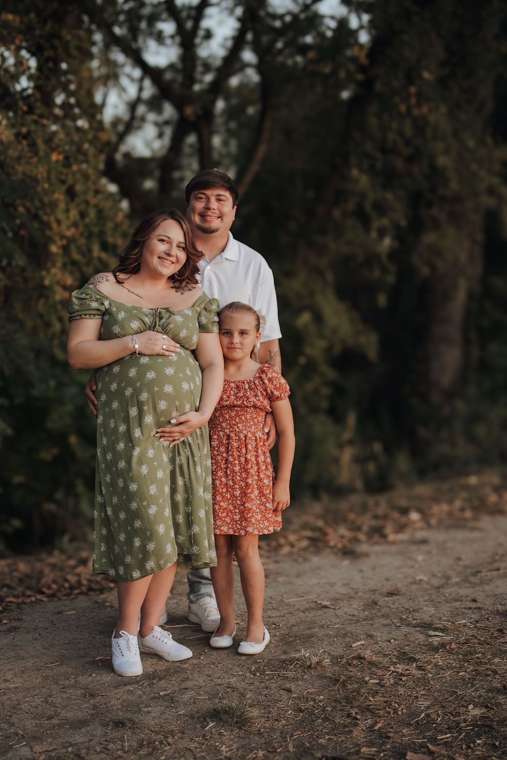 Family stands together outdoors in nature wearing polka dot dress and casual attire for a maternity photo.
