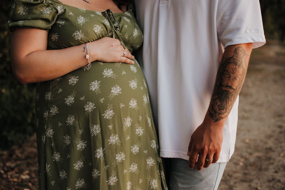 Closeup of a pregnant woman in a green floral dress standing next to someone wearing a white t-shirt with tattoos.