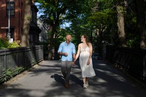 Couple holding hands walking on a tree-lined path in a sunny urban neighborhood, woman in white dress, man in blue shirt.