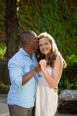 A happy interracial couple shares a romantic kiss outdoors, with the man in blue and woman in white dress smiling.