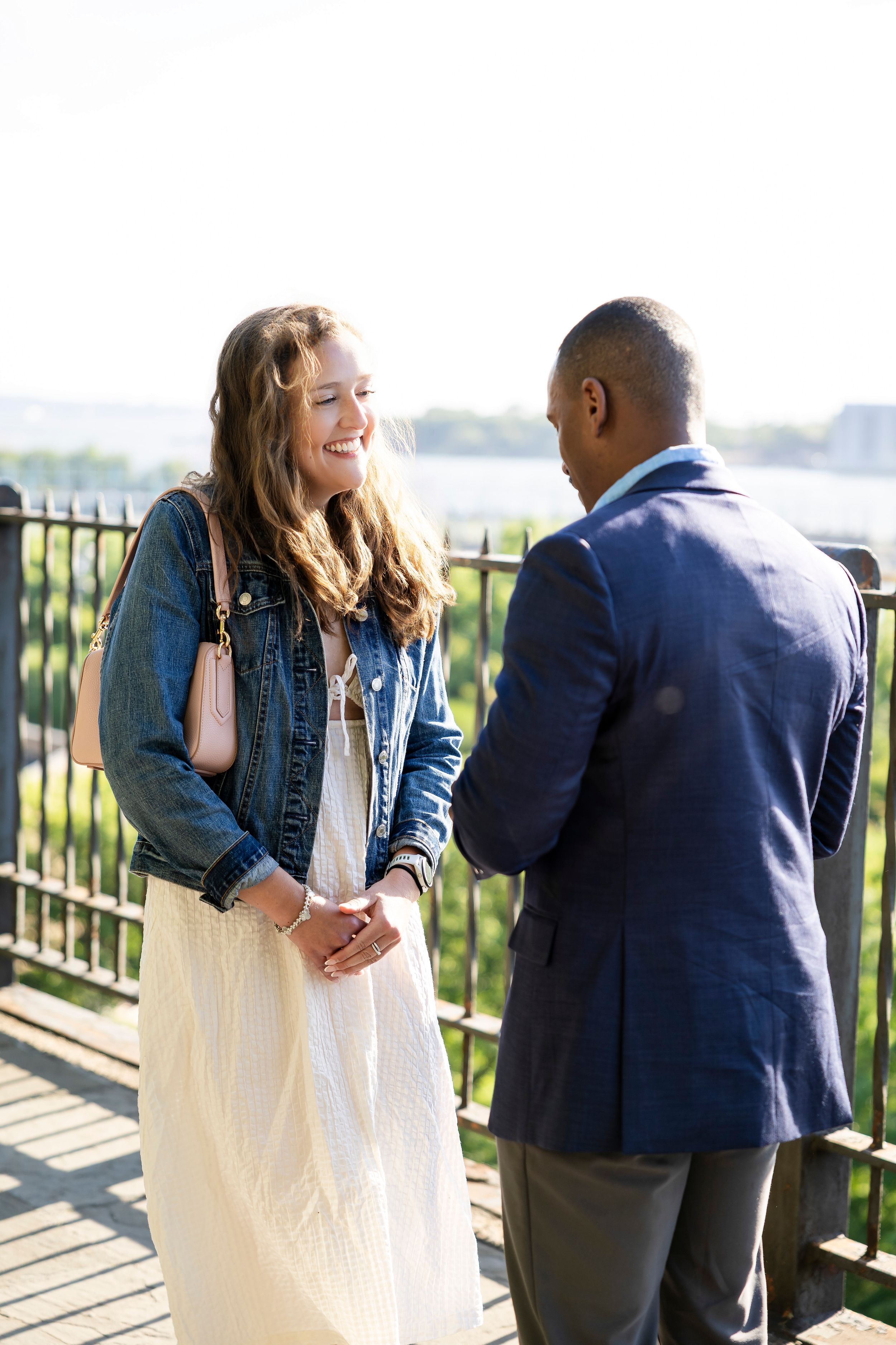 A smiling woman in a denim jacket and white dress talks with a man in a blue blazer outdoors near a railing.