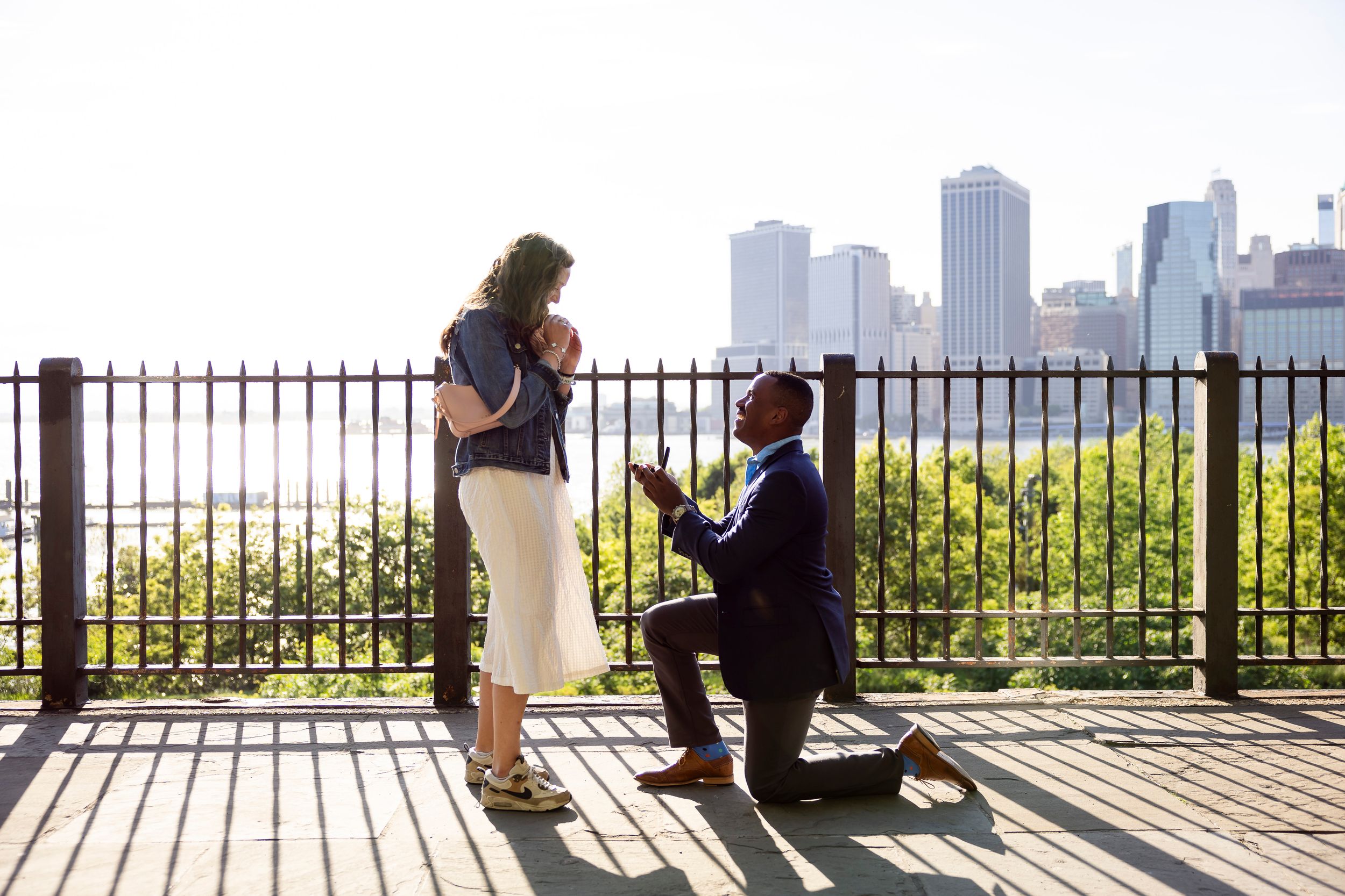 Man proposing to woman on one knee at outdoor overlook with city skyline backdrop on sunny day.