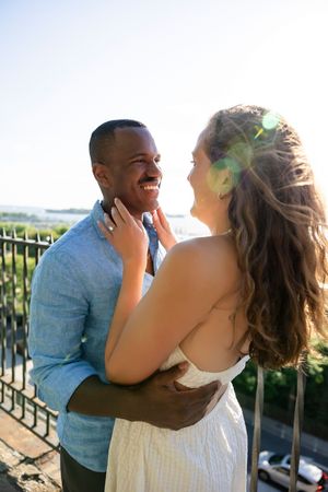 Interracial couple embracing outdoors, smiling at each other in warm sunlight near a railing.