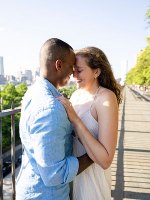 Interracial couple sharing a tender moment on a bridge with NYC skyline in the background on a sunny day.