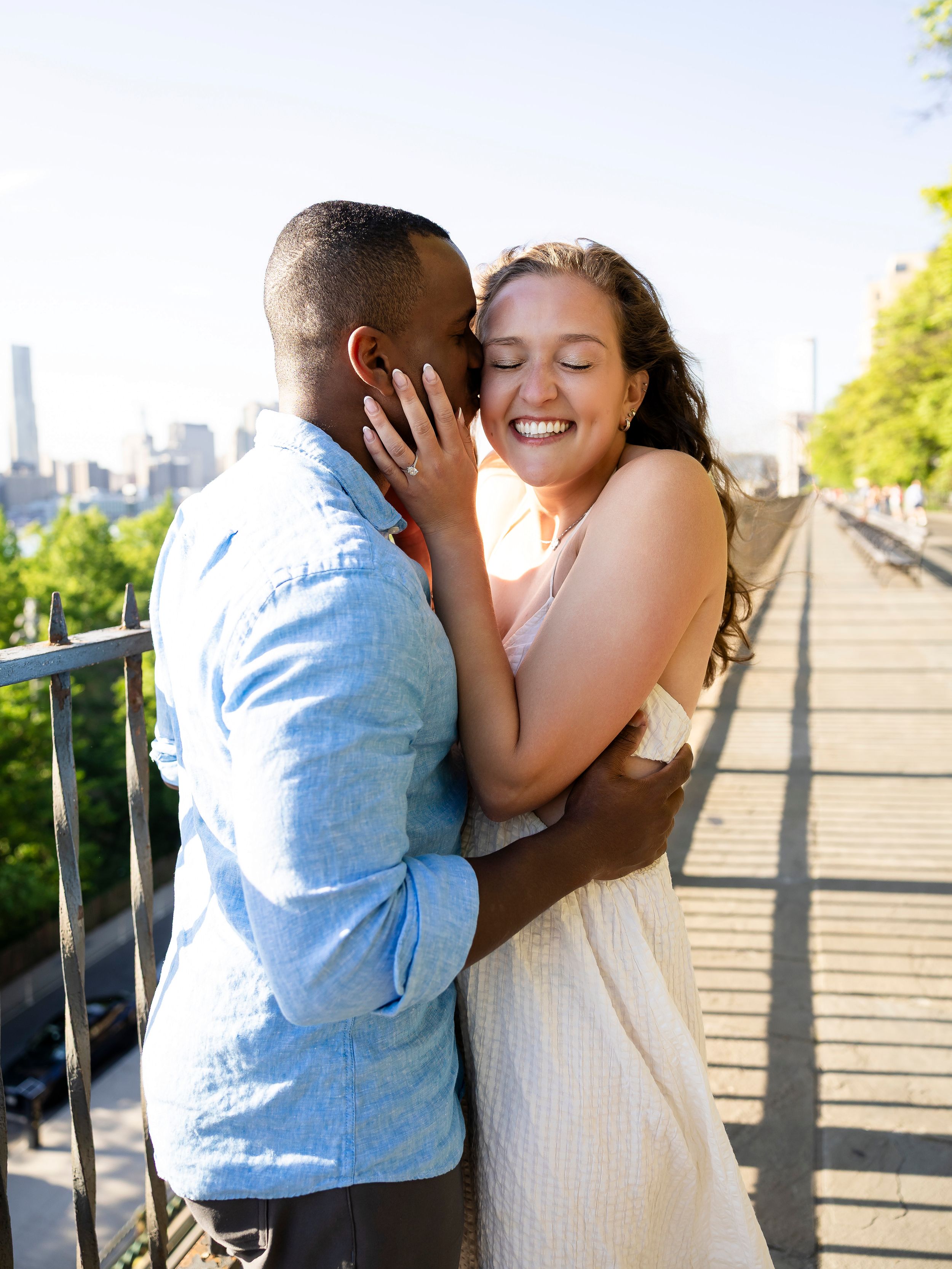 Joyful interracial couple embracing and laughing together on a sunny NYC bridge with city skyline views.