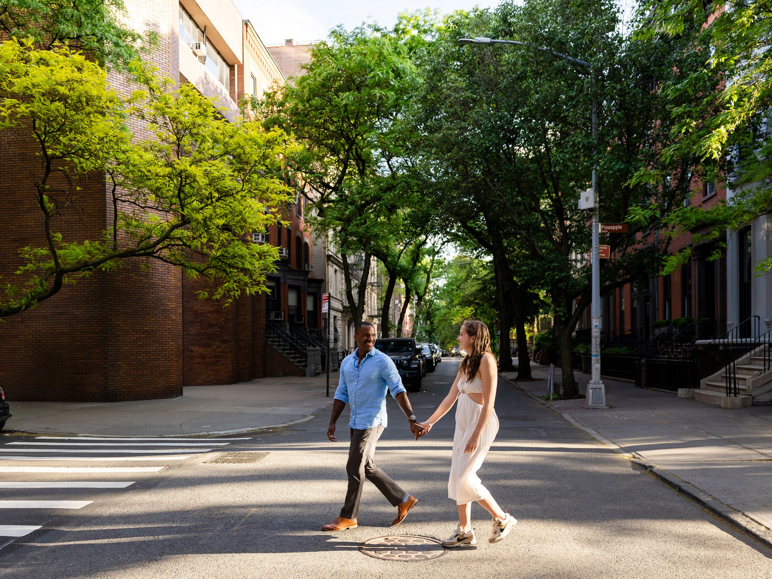 A couple holds hands while crossing a sunny urban street lined with lush green trees and brick buildings.