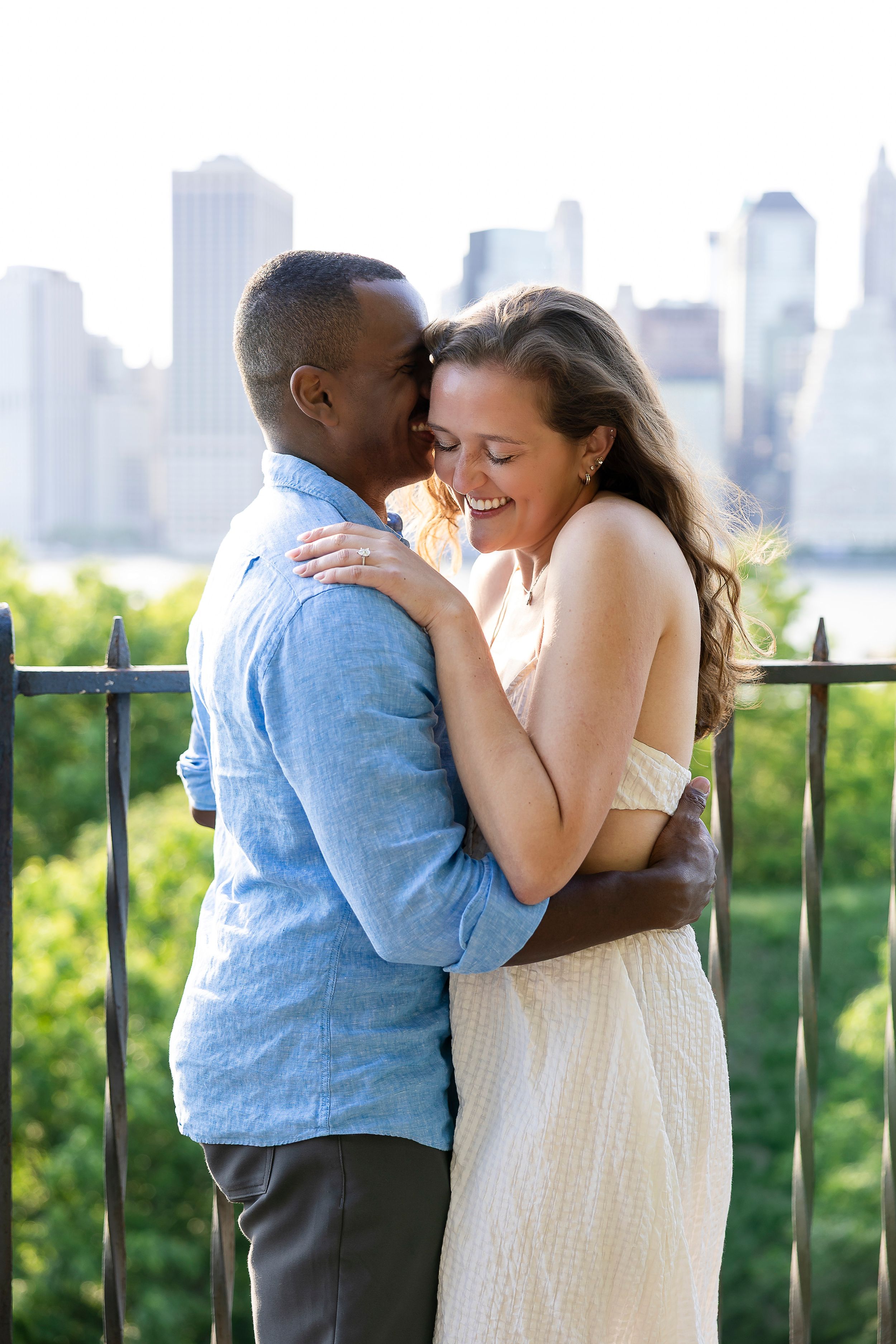 Happy interracial couple embracing and laughing together outdoors with city skyline and greenery in background.
