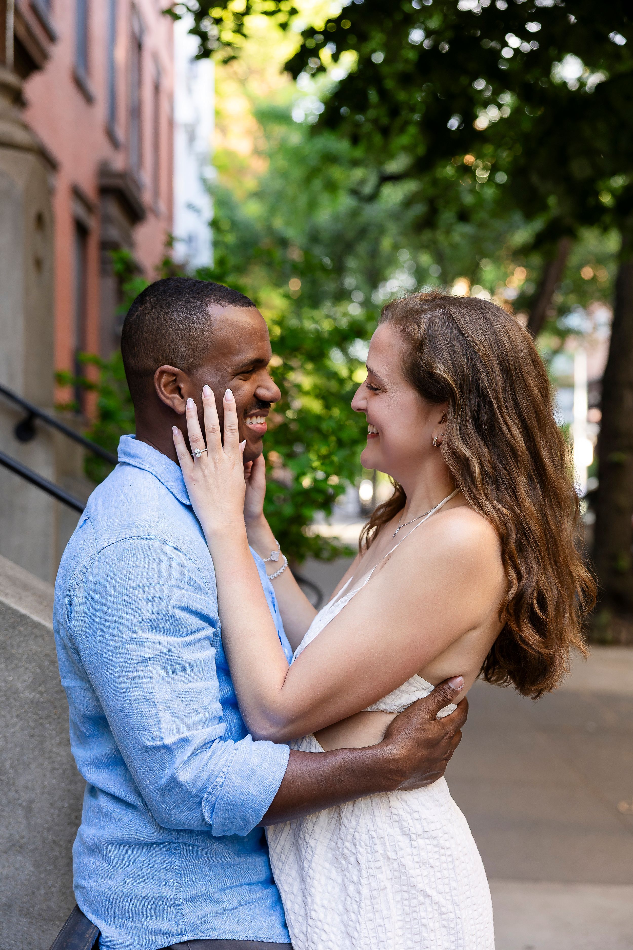 A joyful interracial couple embraces and smiles at each other on a tree-lined urban street.