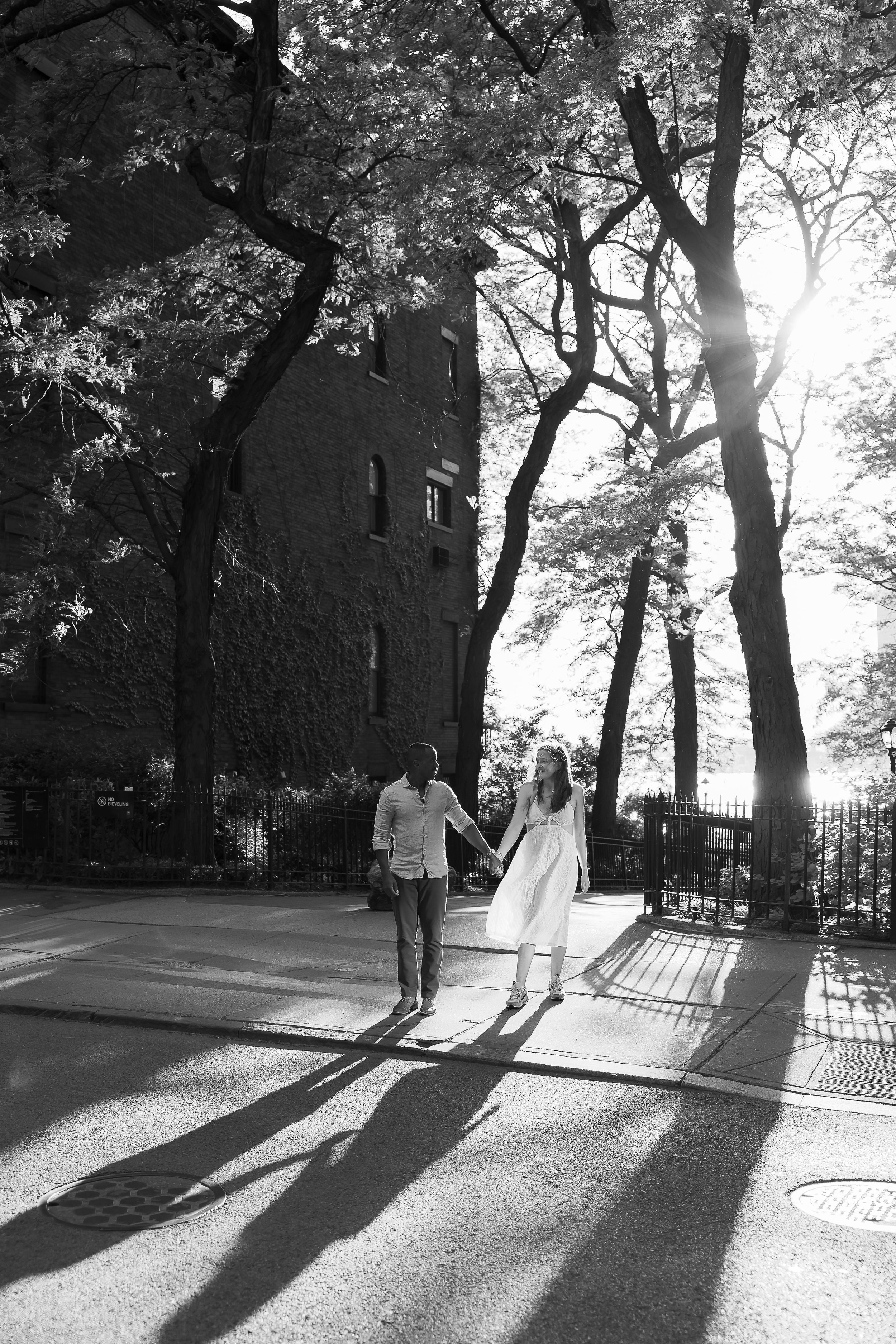 Black and white photo of a couple holding hands walking on a sunny tree-lined sidewalk near a brick building.