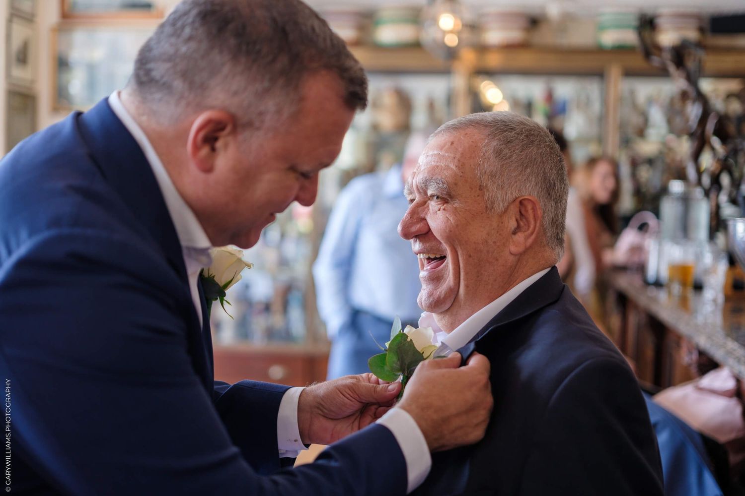 Two well-dressed men in suits share a joyful moment while one adjusts the other's boutonniere at an indoor event.