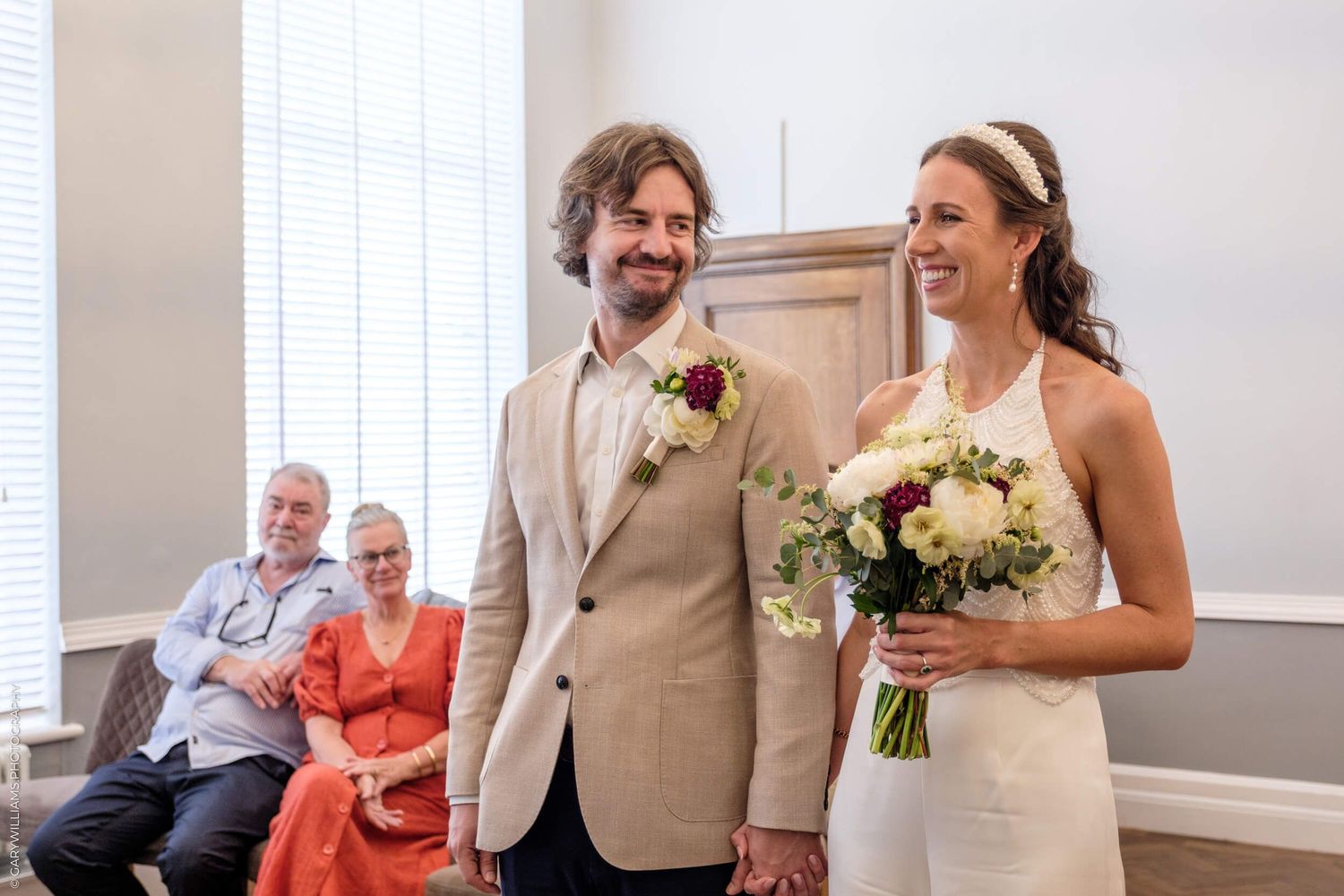 A joyful couple laughs together during their wedding ceremony while elderly guests watch from chairs in the background.