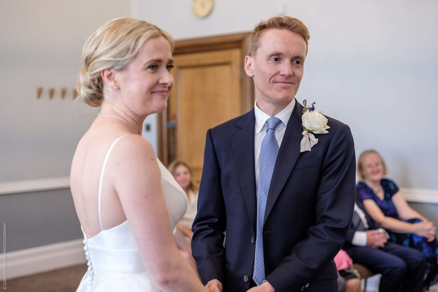 A bride in a white gown and groom in a blue tie share a moment during their wedding ceremony.