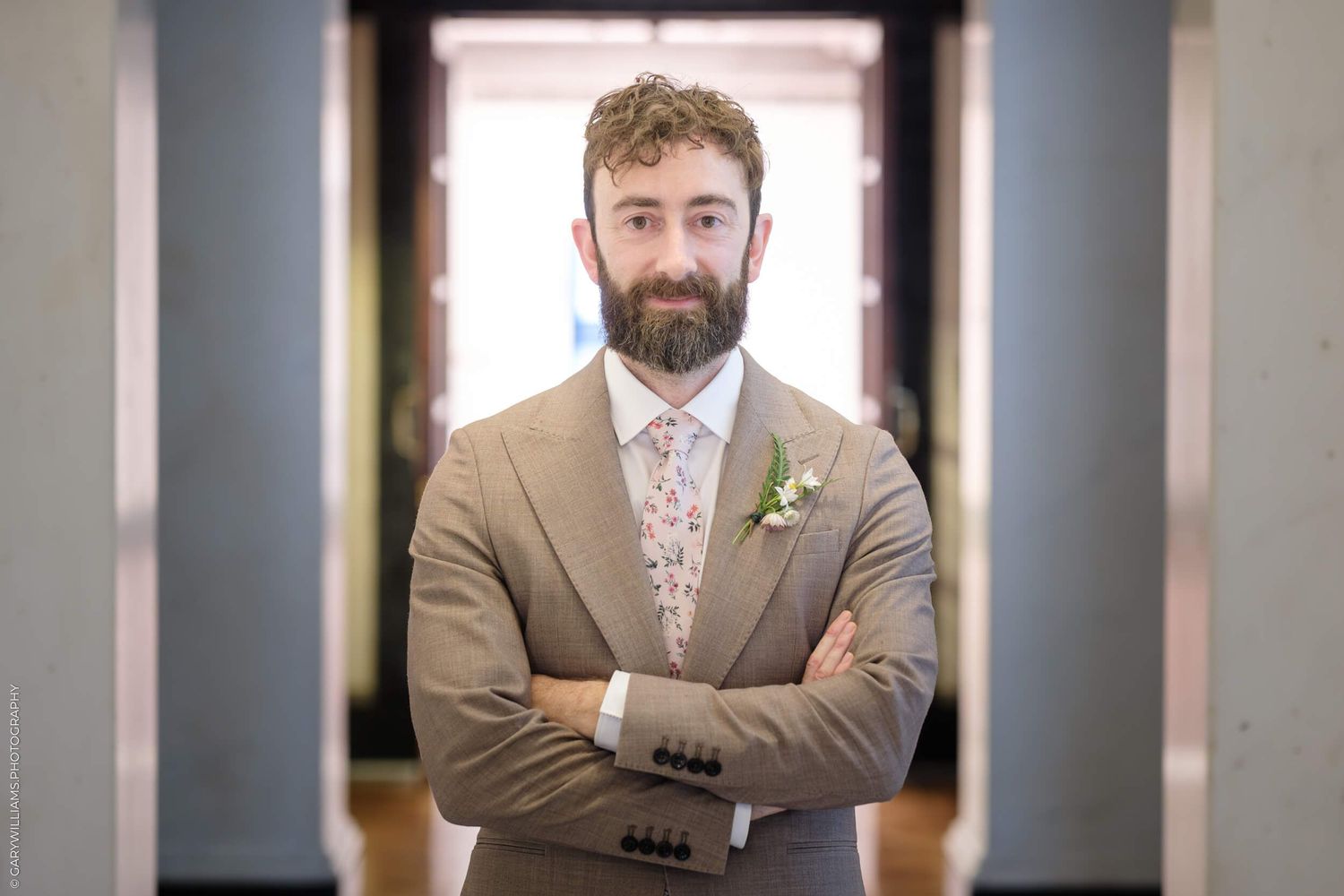 A groom wearing a tan suit and floral tie stands with arms crossed in a hallway during a wedding.