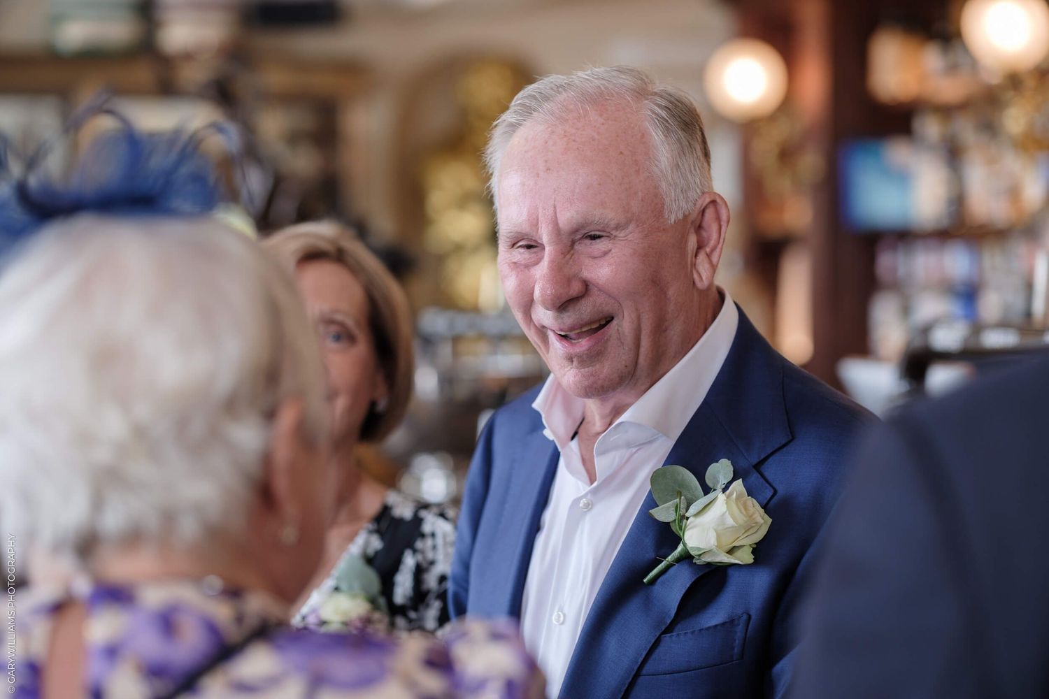 An elegantly dressed man in a navy suit and white boutonniere smiles during a social gathering in a warm indoor setting.