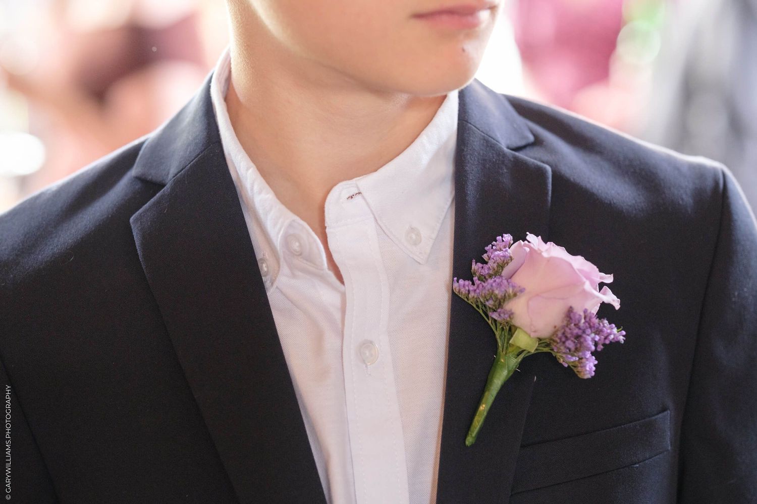 Groomsmen wearing black suits with lilac boutonnieres stand in a row at a formal wedding ceremony.