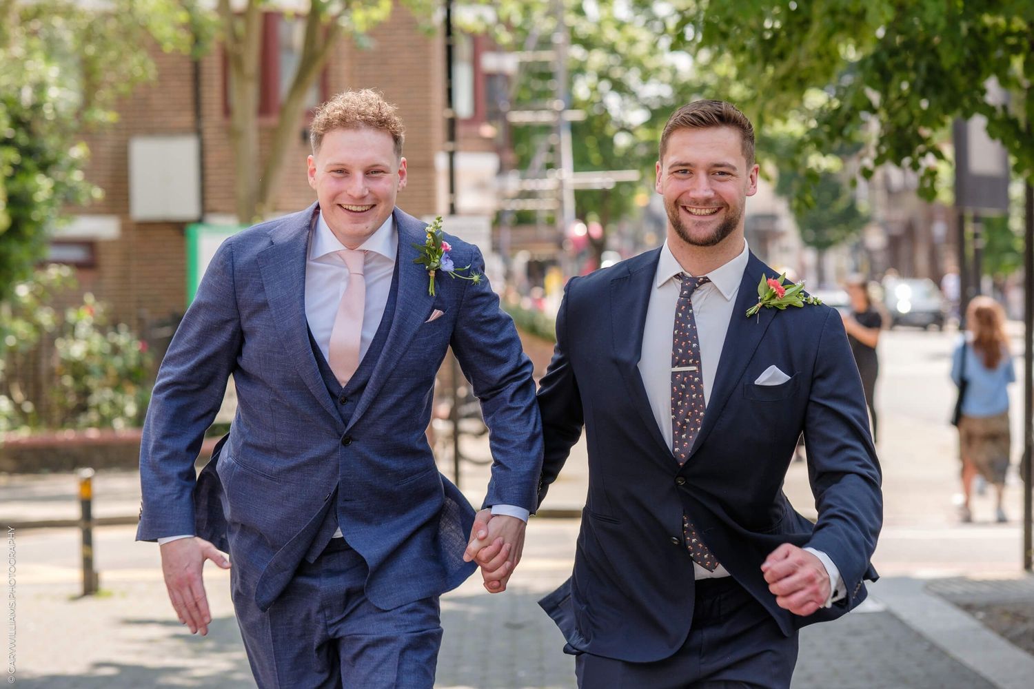Two people wearing navy blue suits run joyfully down a tree-lined city street on a sunny day.