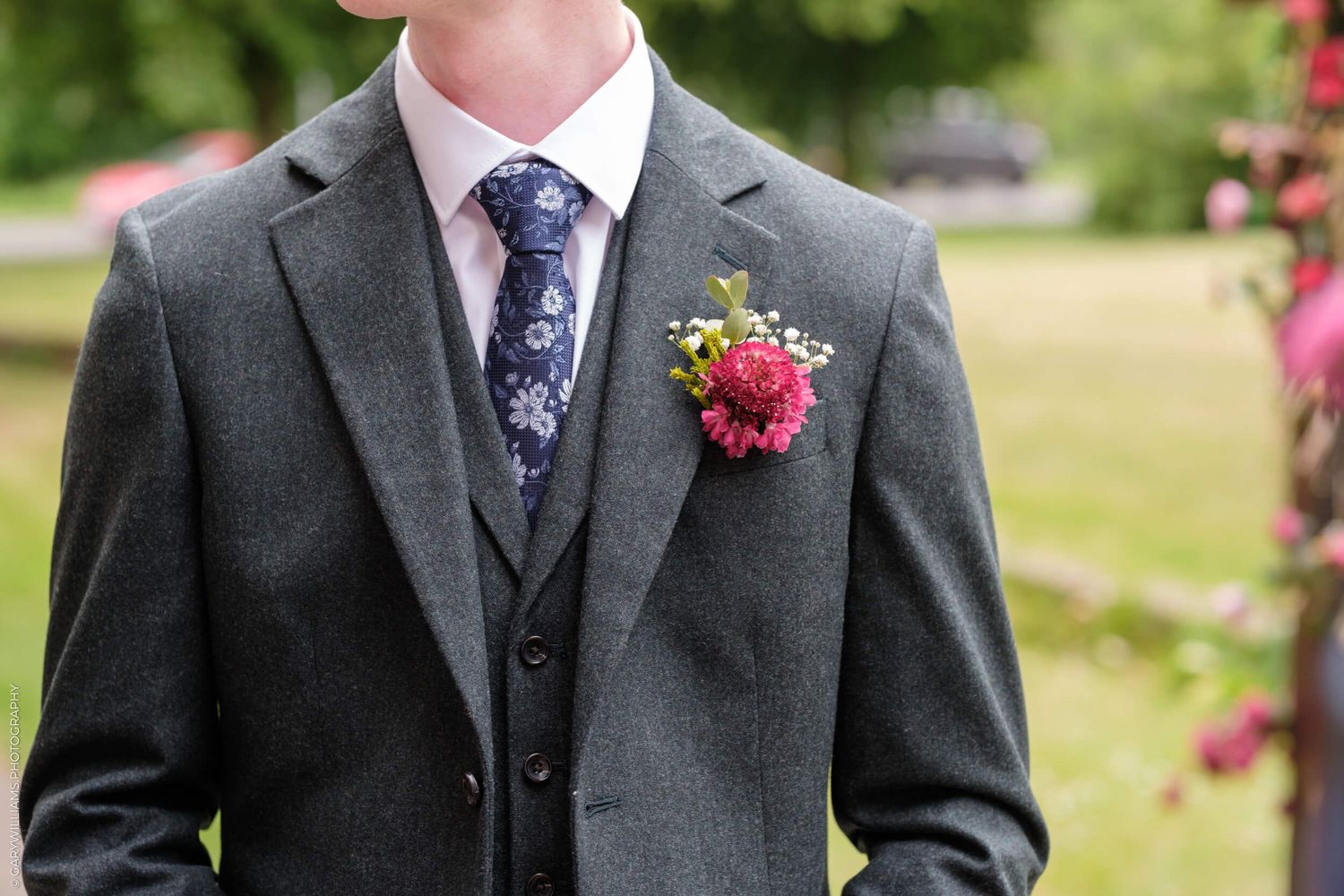 Close-up view of a charcoal gray suit jacket with a purple floral tie and pink boutonniere at a wedding.