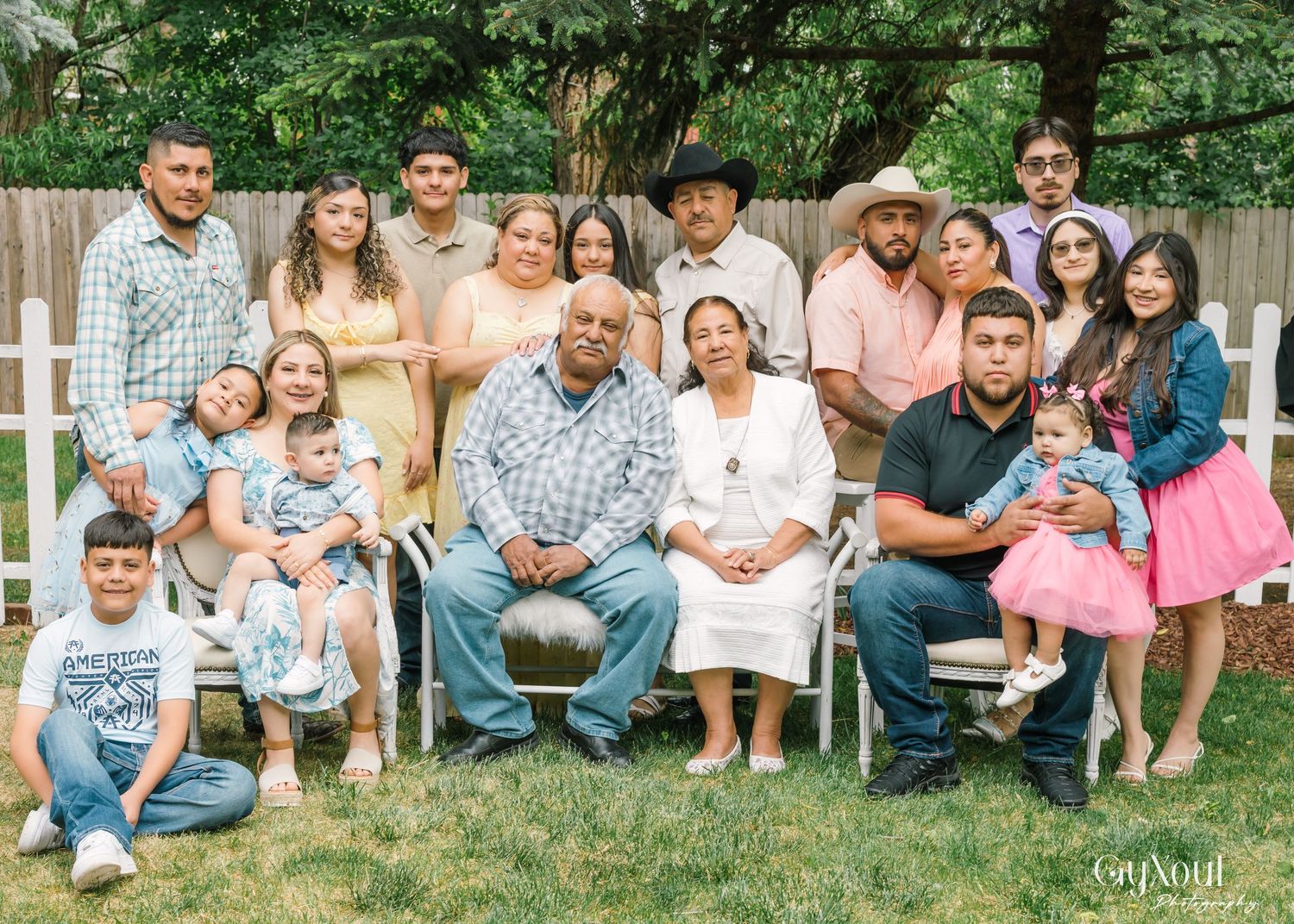 A large multi-generational family poses together outdoors in a backyard setting with white fence and greenery.