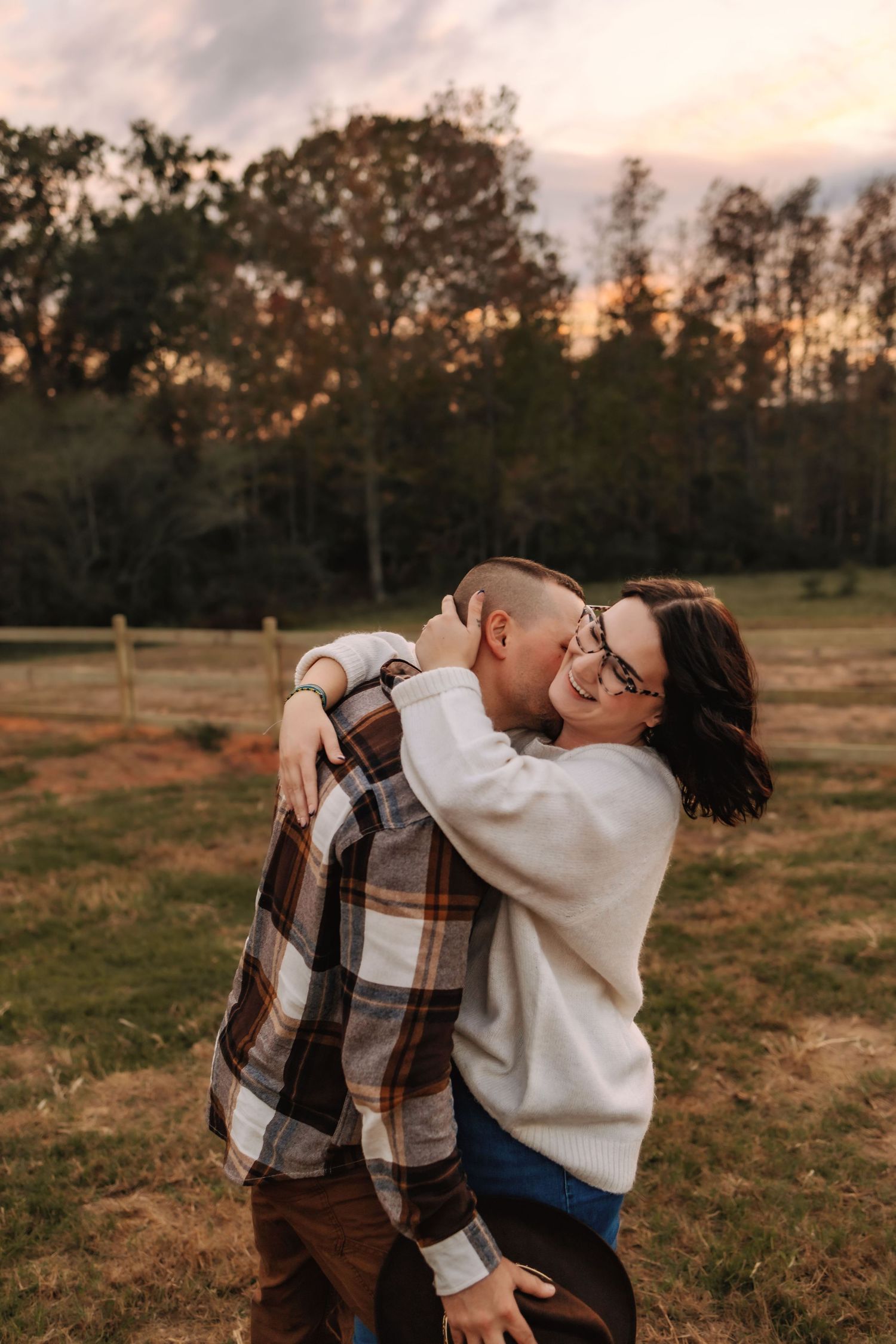 A couple embraces in a field at sunset with trees silhouetted in the background.