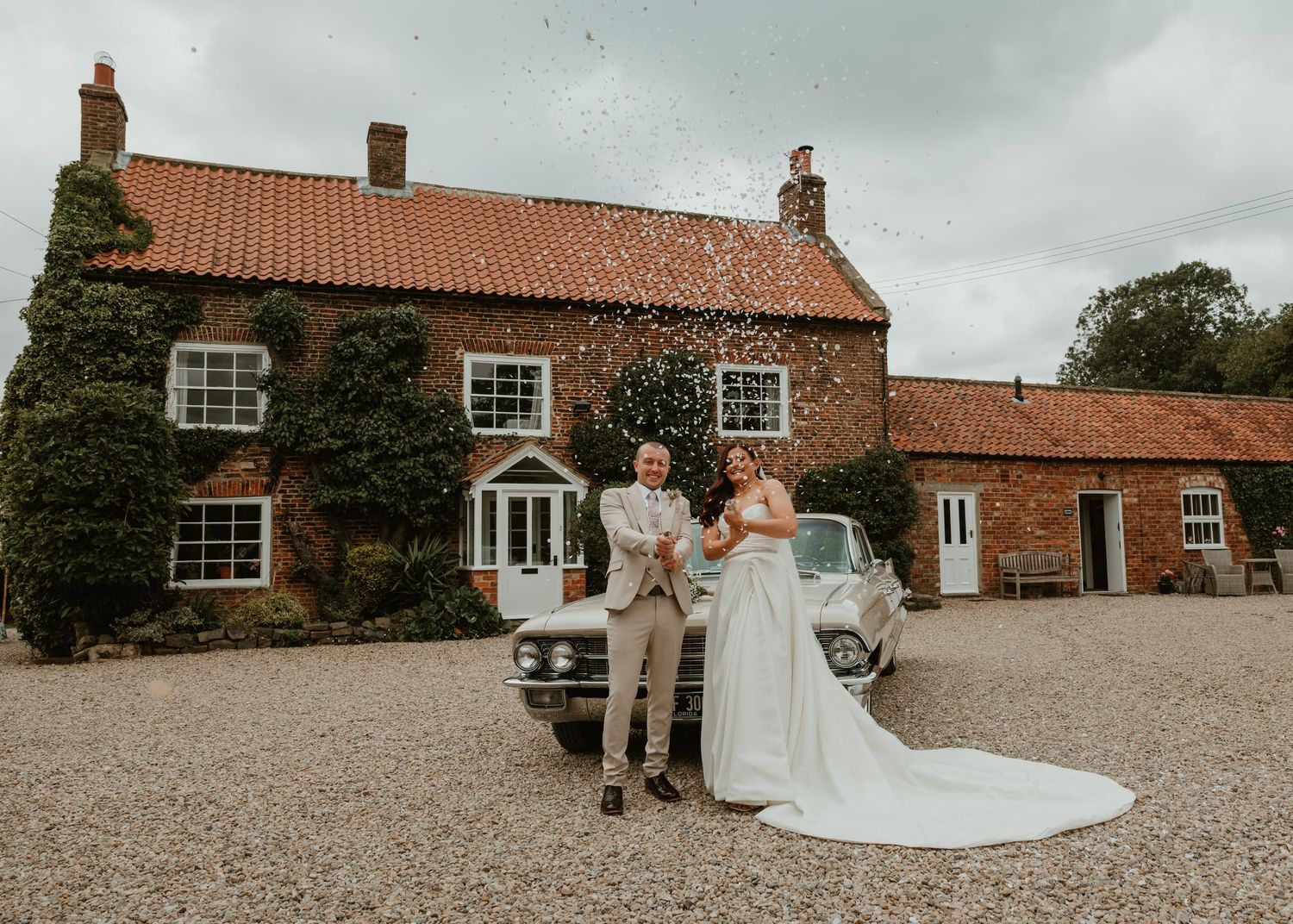Row of red brick buildings at rustic countryside wedding venue with classic cars parked outside.