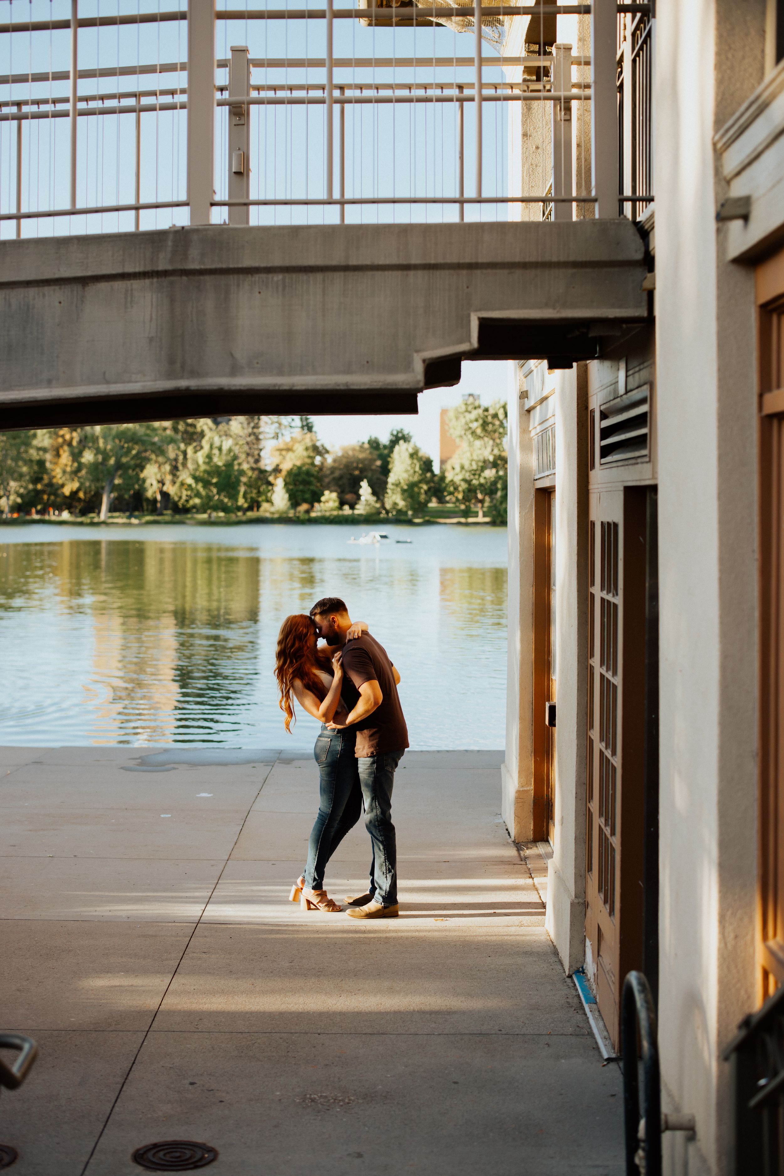 A couple sharing an intimate moment on a riverside walkway beneath a bridge during golden hour.