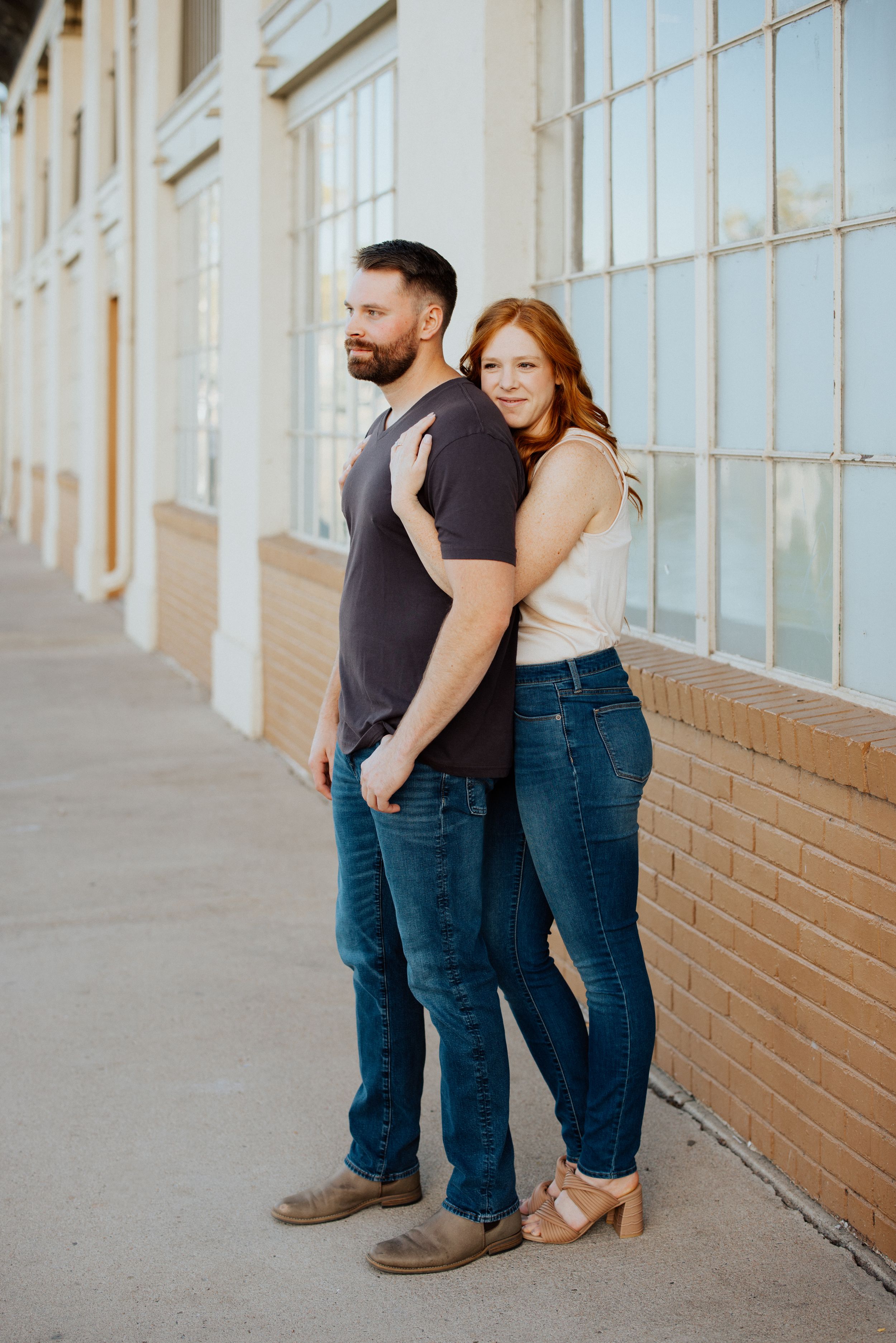A fashionable couple in casual attire stands side by side against a brick and window wall on an urban sidewalk.