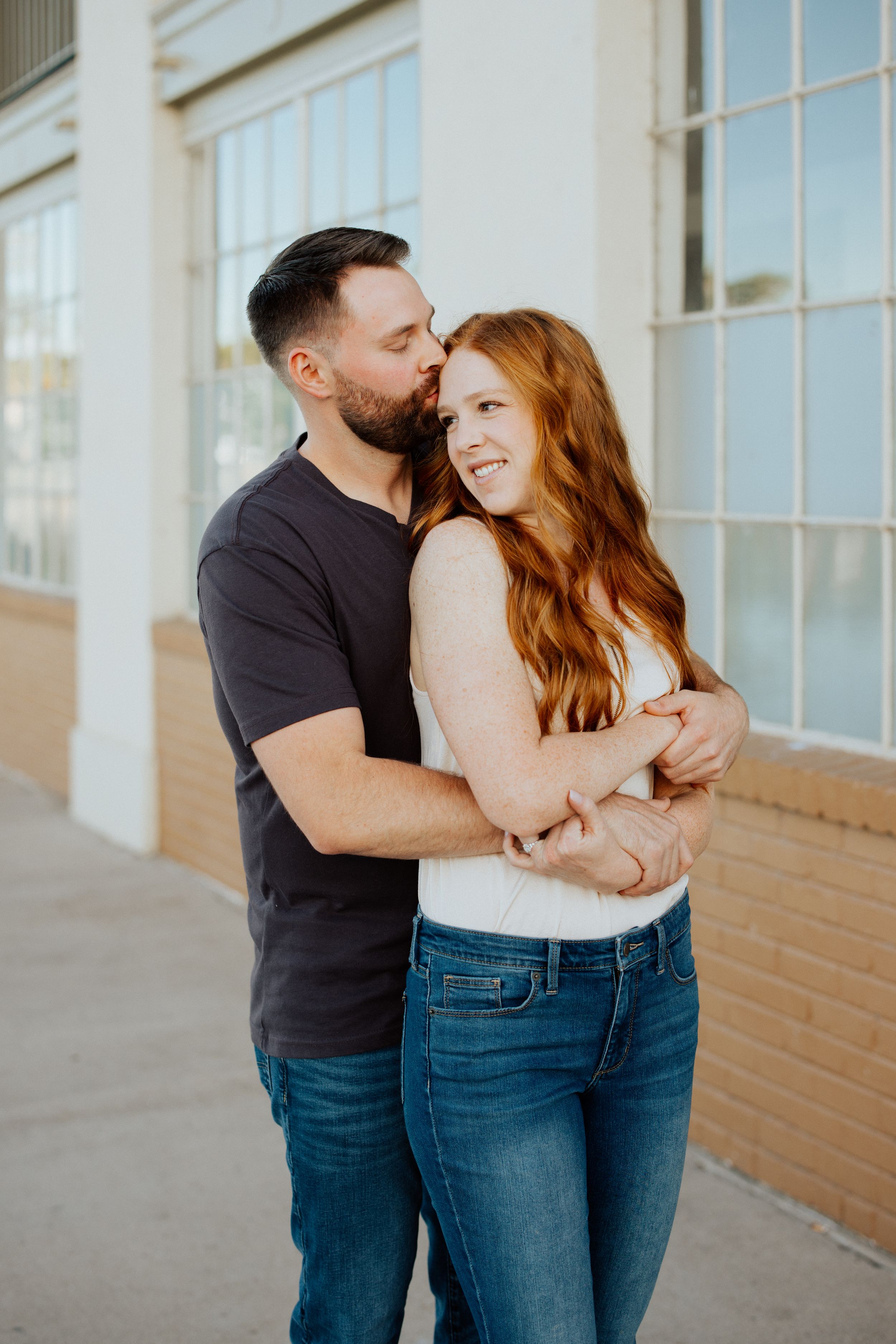 Affectionate couple sharing a tender moment outdoors against a brick building in warm sunlight.