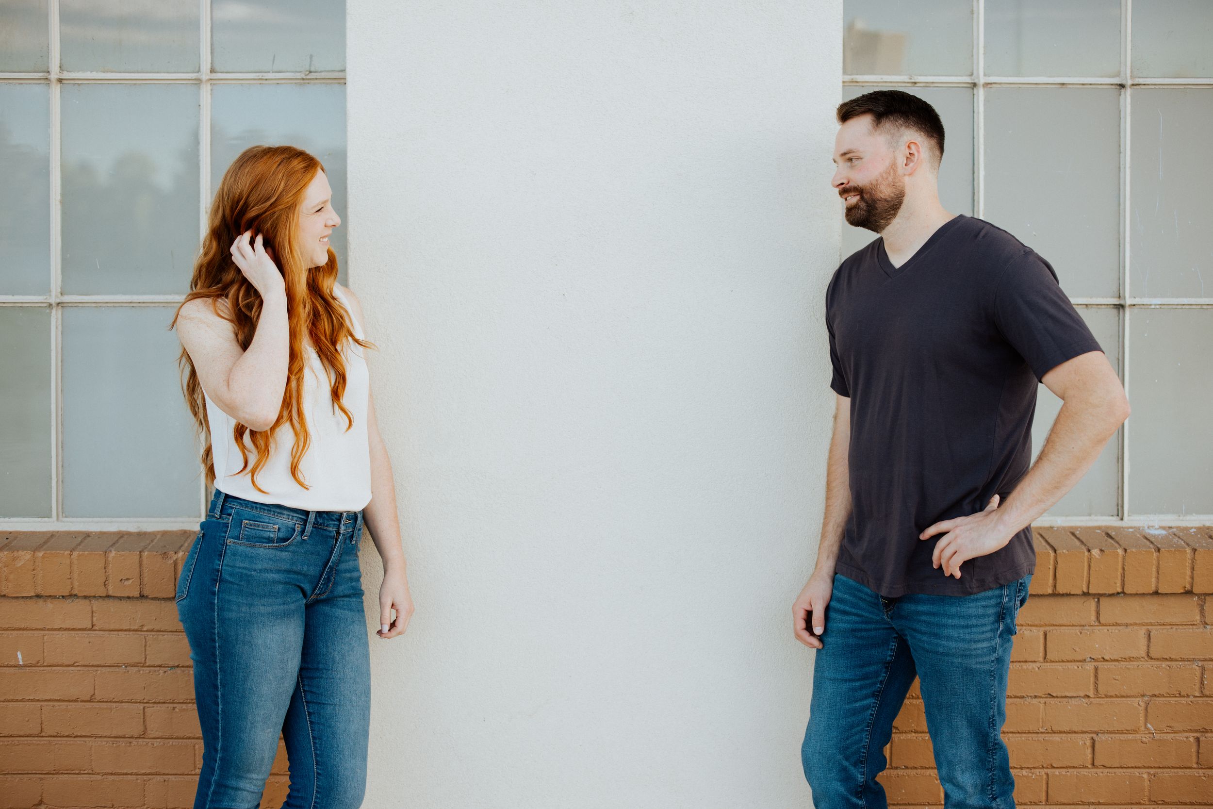 Woman in white top and jeans laughs while leaning against wall with brown bricks and frosted glass windows.