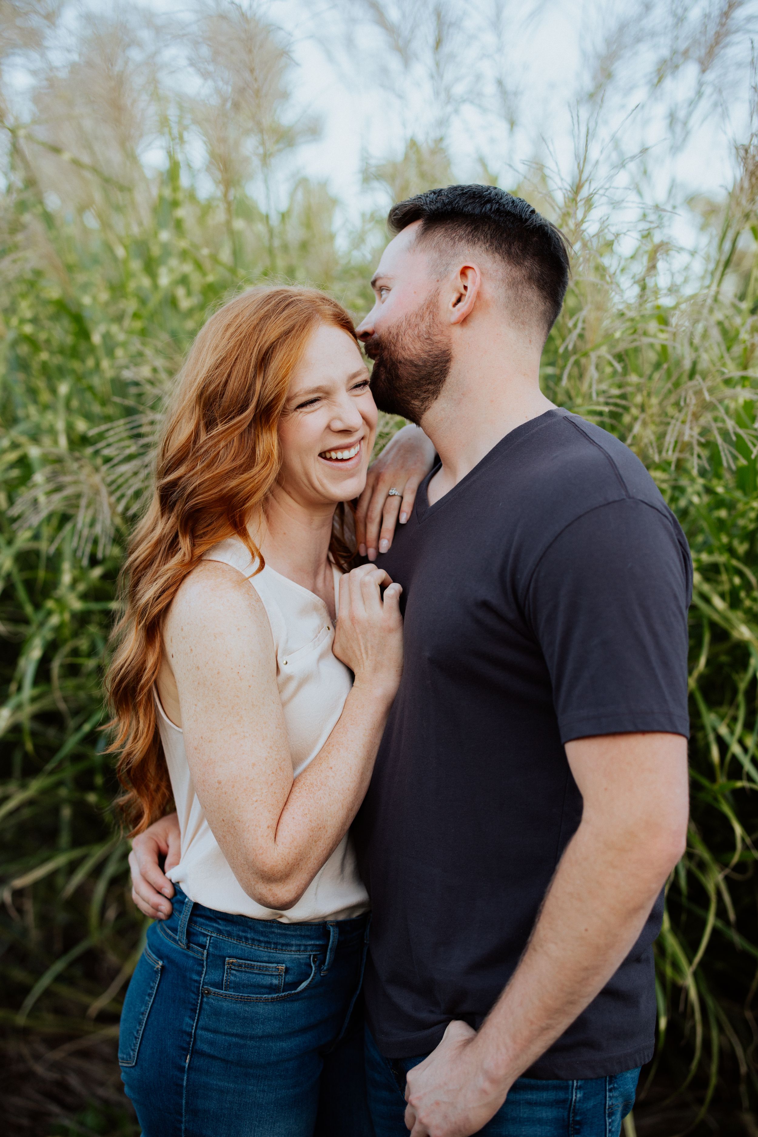 Couple shares intimate laughing moments together against tall green grass background in casual outdoor photoshoot.