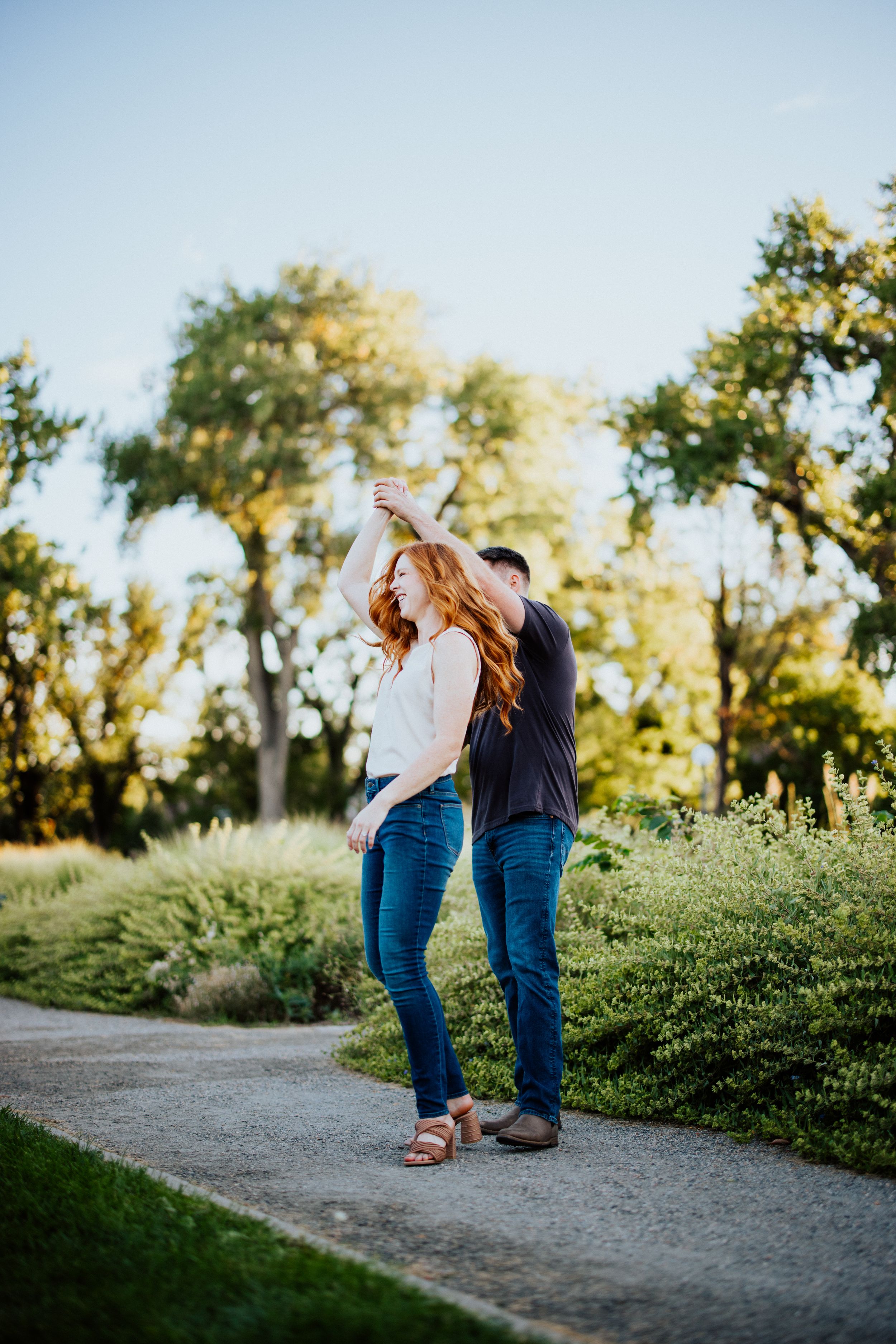 A romantic couple embraces on a dirt path surrounded by garden greenery and trees in warm sunset lighting.