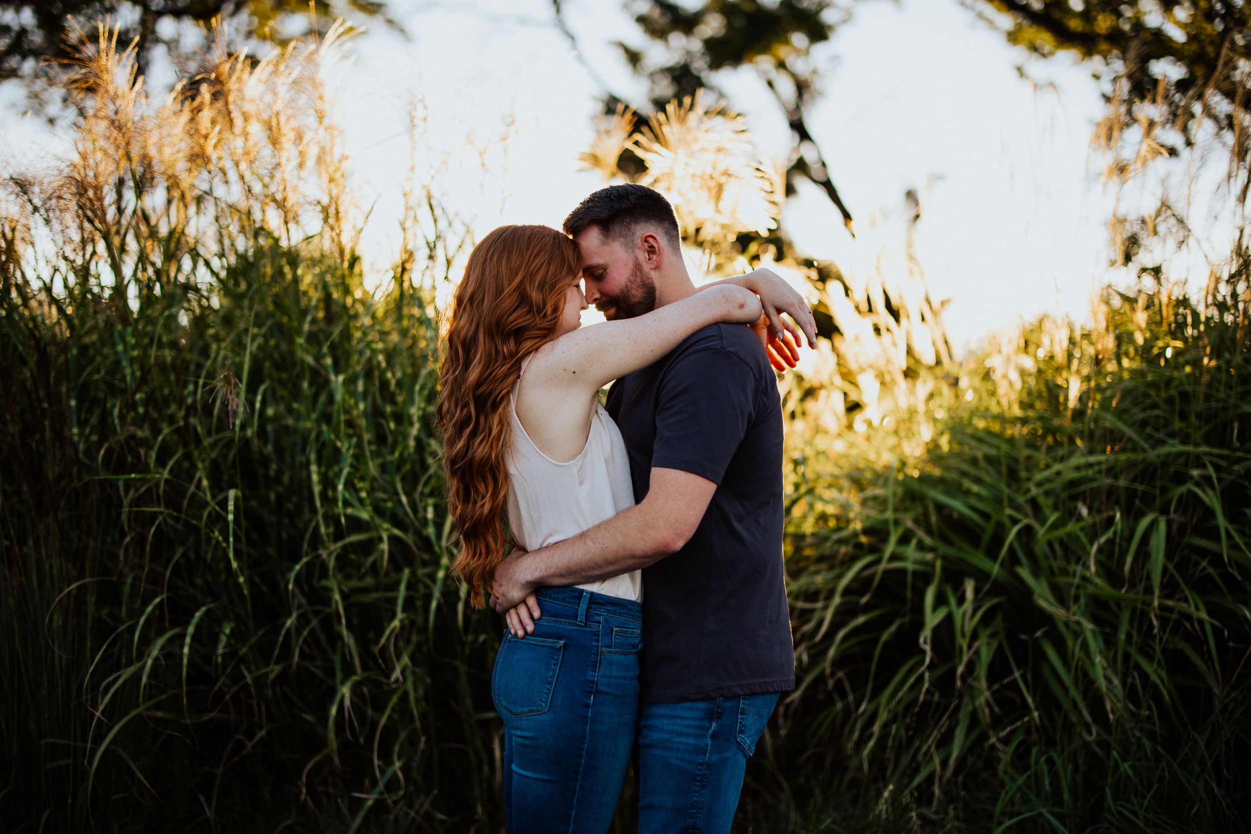 A couple embraces lovingly in front of a golden sunlit cornfield during a sunset photoshoot.