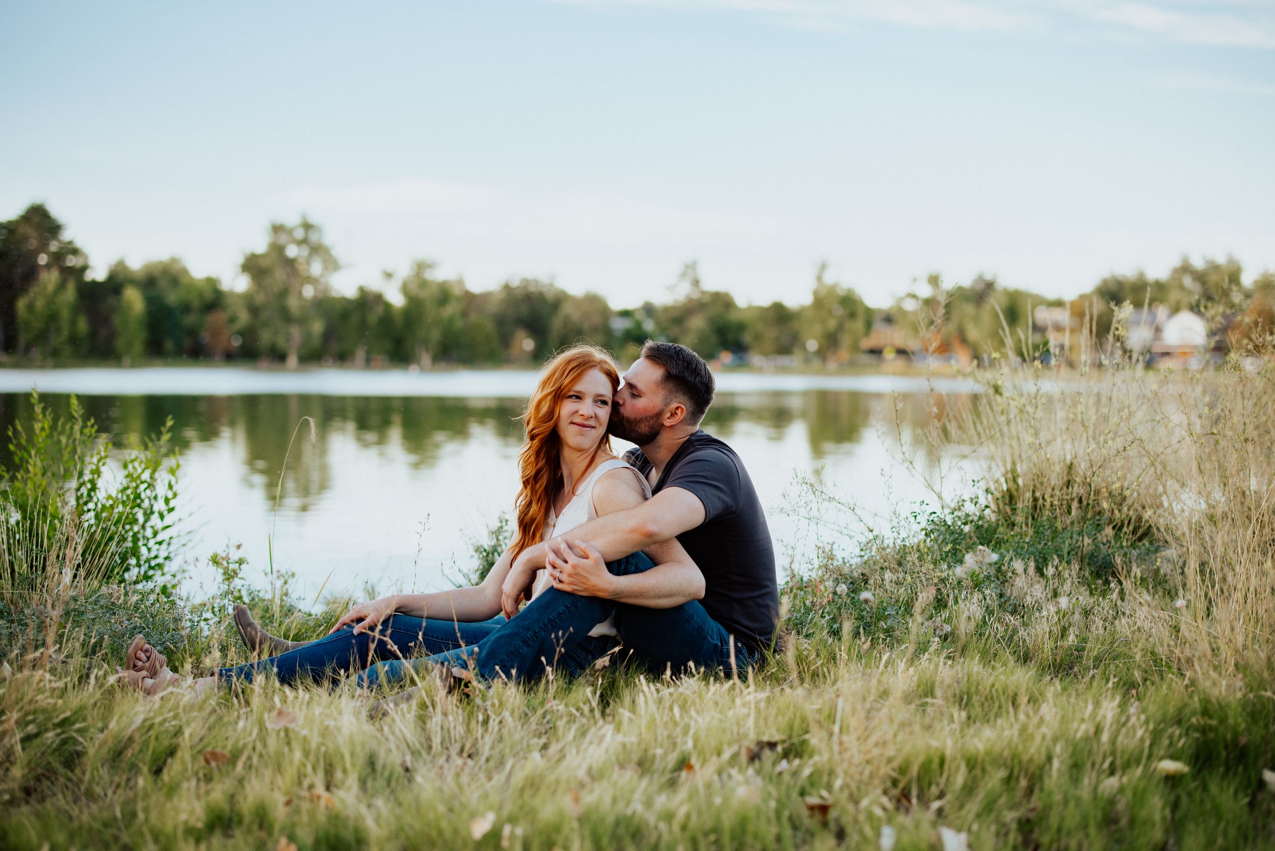 A couple share an intimate moment together in a grassy meadow near a serene lake at sunset.