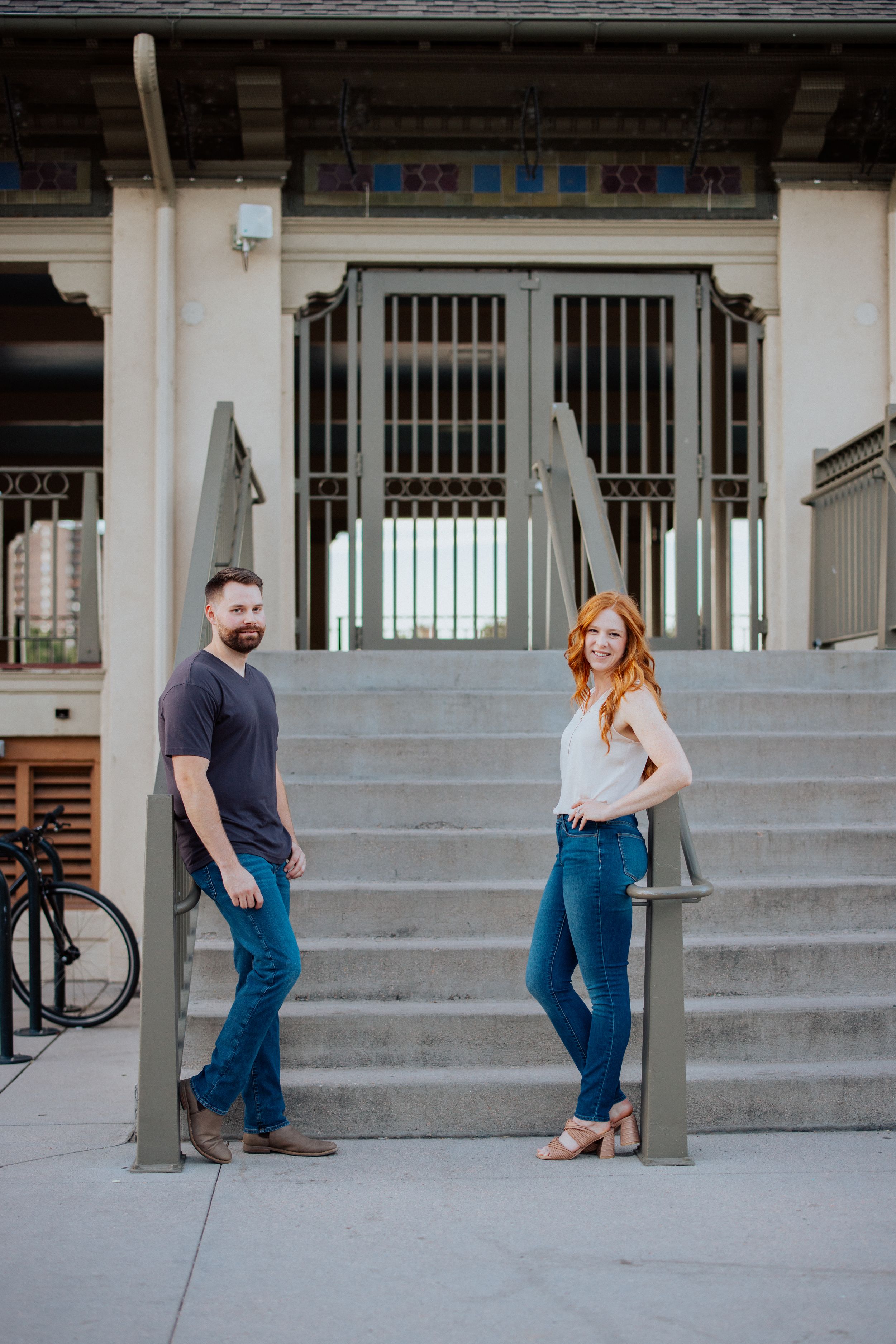 Couple poses playfully on concrete steps outside a building during casual outdoor photo session.