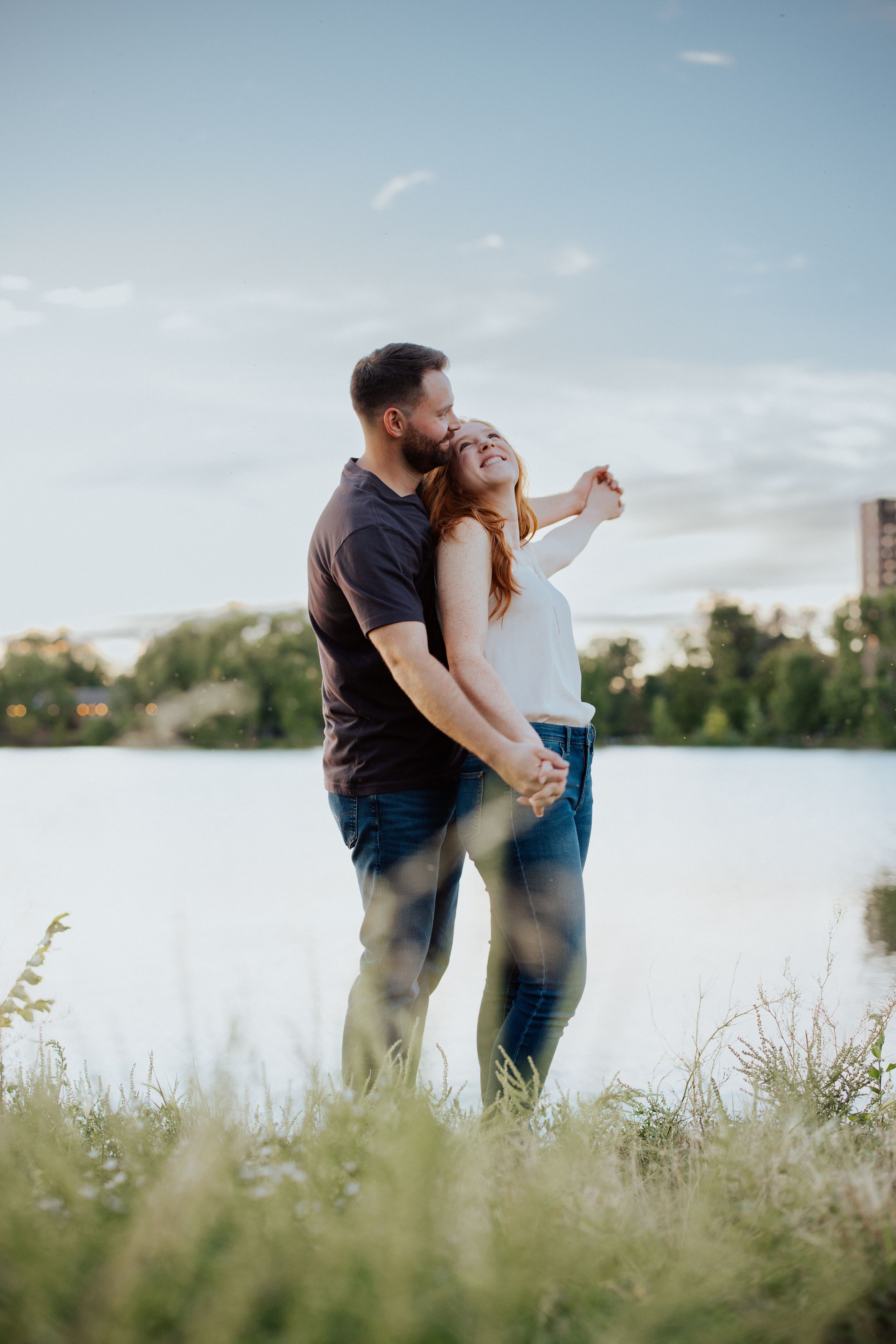 A couple shares a loving moment by a tranquil lake during sunset with wild grass in the foreground.