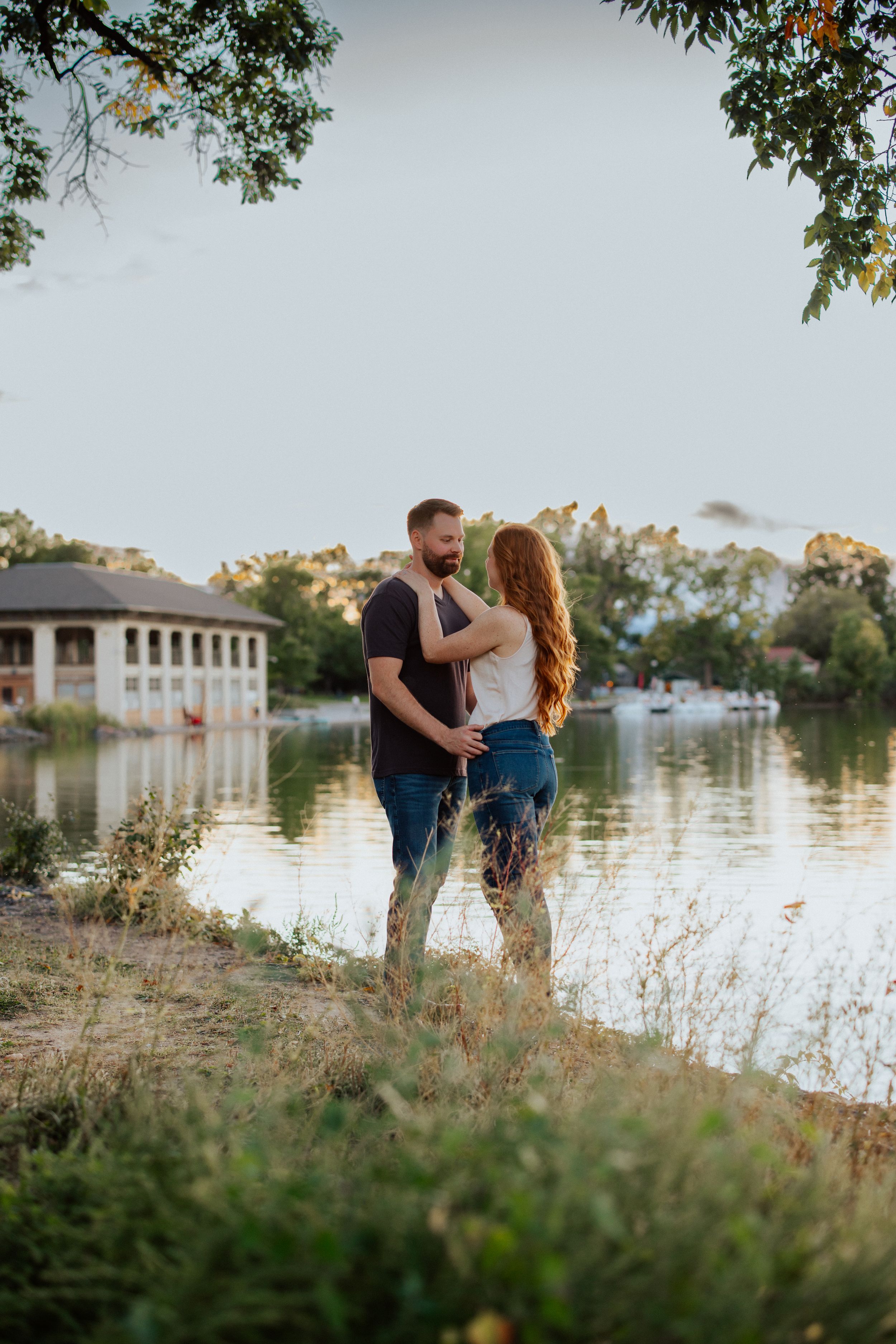 A romantic couple embraces by a serene lake at sunset with a gazebo in the background and wildflowers in the foreground.