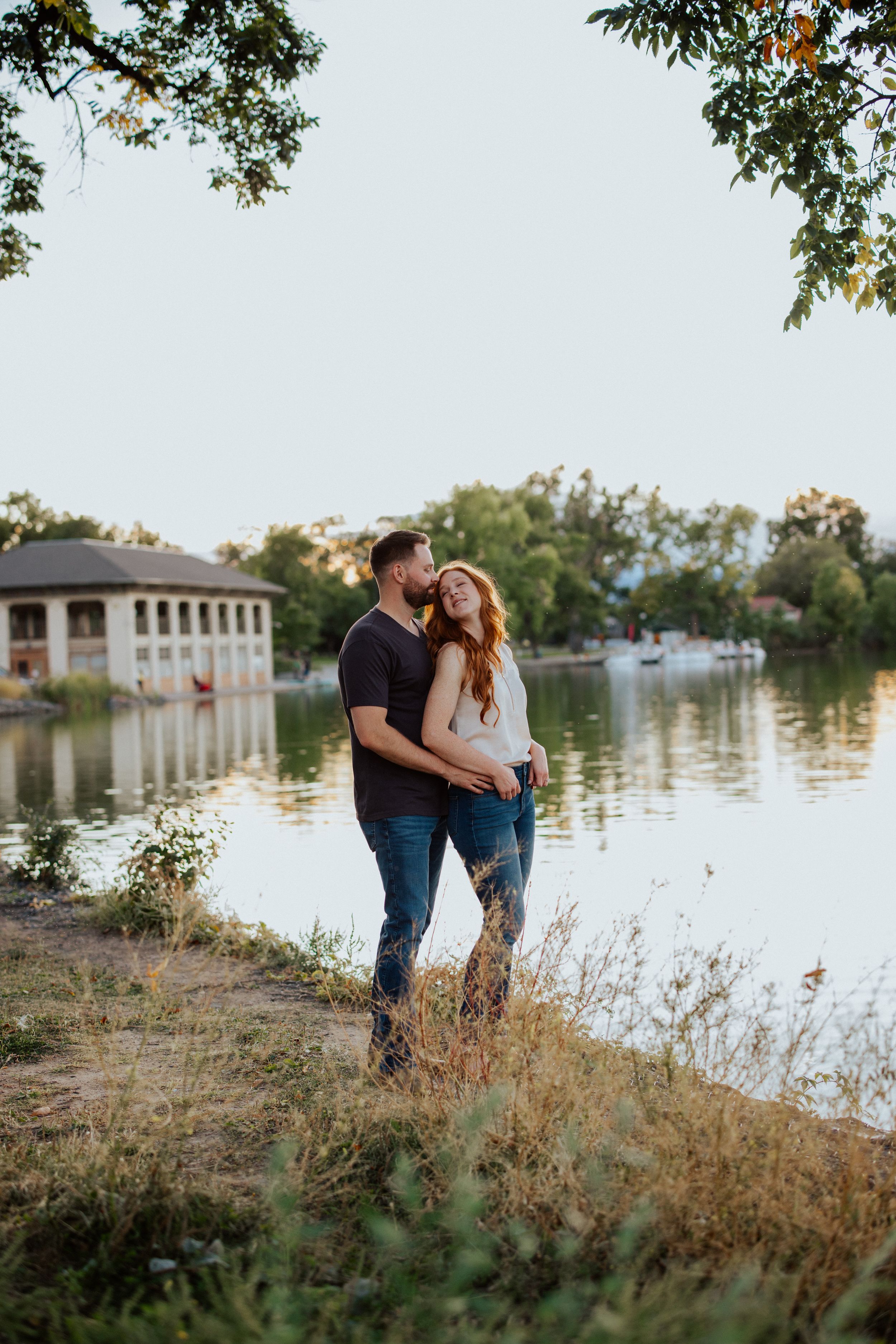 Romantic couple embracing by a scenic lakeside with white columns and trees in the background.